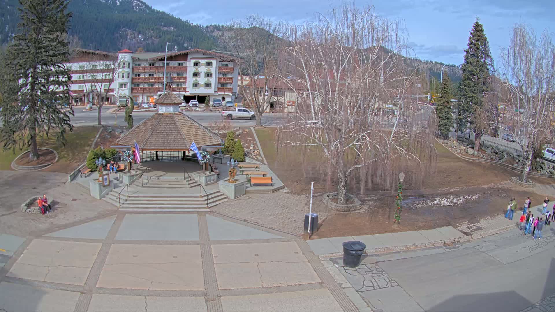 A park with a gazebo, benches, and a few people, situated next to a street and a multi-story building, all under a sunny, mostly clear sky.
