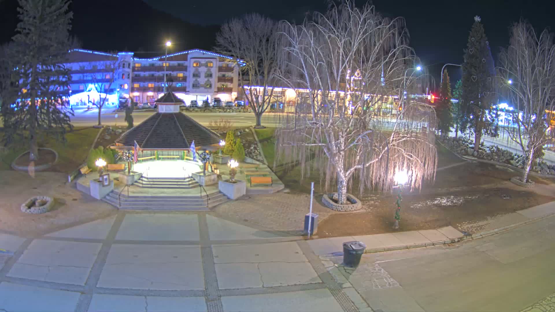 A nighttime view of a town square with a gazebo, bare trees, and a multi-story building illuminated with lights.