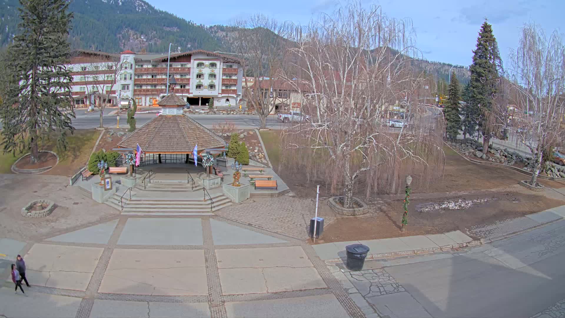 A paved town square with a gazebo, benches, and bare trees, under a partly sunny sky, is situated in front of a multi-story building and mountains in the distance.