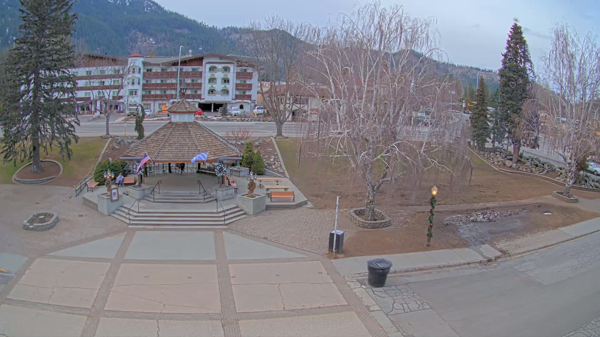 An overcast day reveals a town square with a gazebo, benches, and bare trees, situated near a multi-story building and mountains in the background.