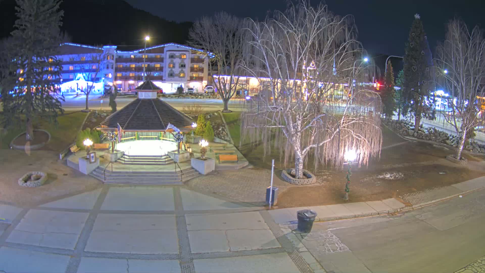 A nighttime view of a town square featuring a gazebo, a decorated Christmas tree, and a multi-story hotel building, all illuminated under clear skies.