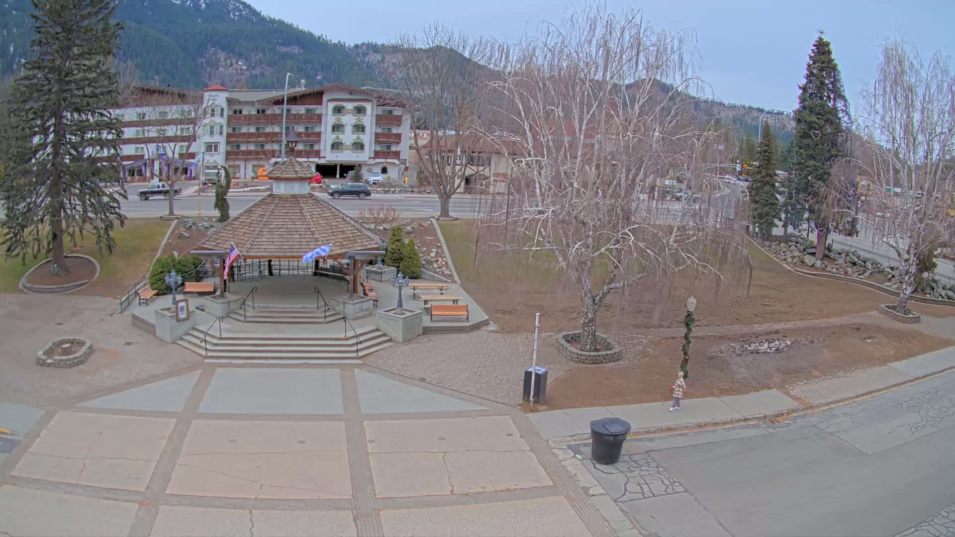 A wooden gazebo with steps sits in a town square, bordered by a street and buildings, under a mostly cloudy sky.