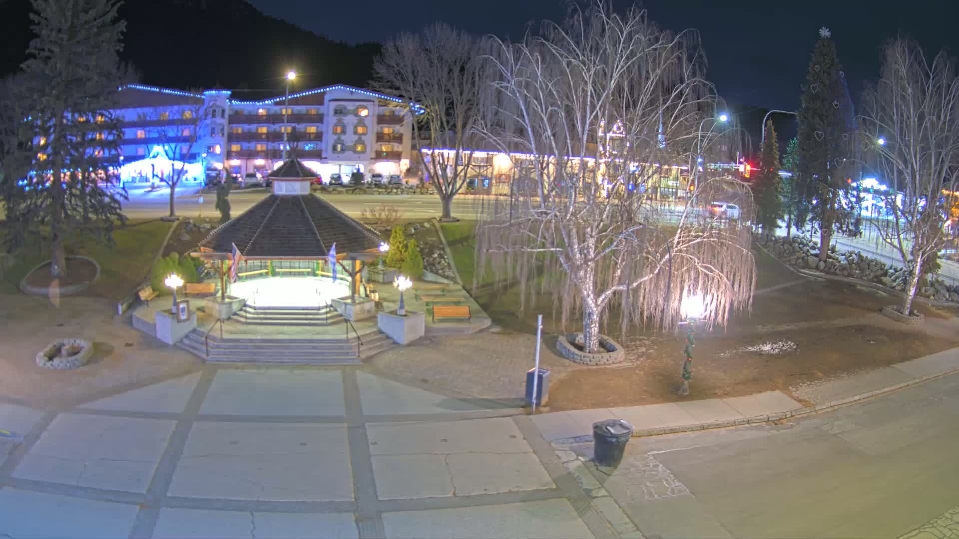 A nighttime view of a town square with a gazebo, a multi-story hotel in the background, bare trees, and a decorated Christmas tree.