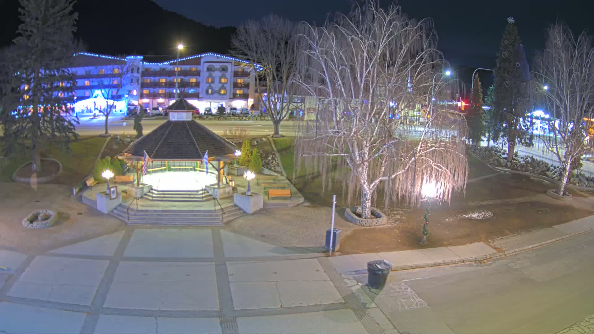 A nighttime view of a town square with a gazebo, bare trees, a decorated Christmas tree, and a hotel in the background under clear skies.