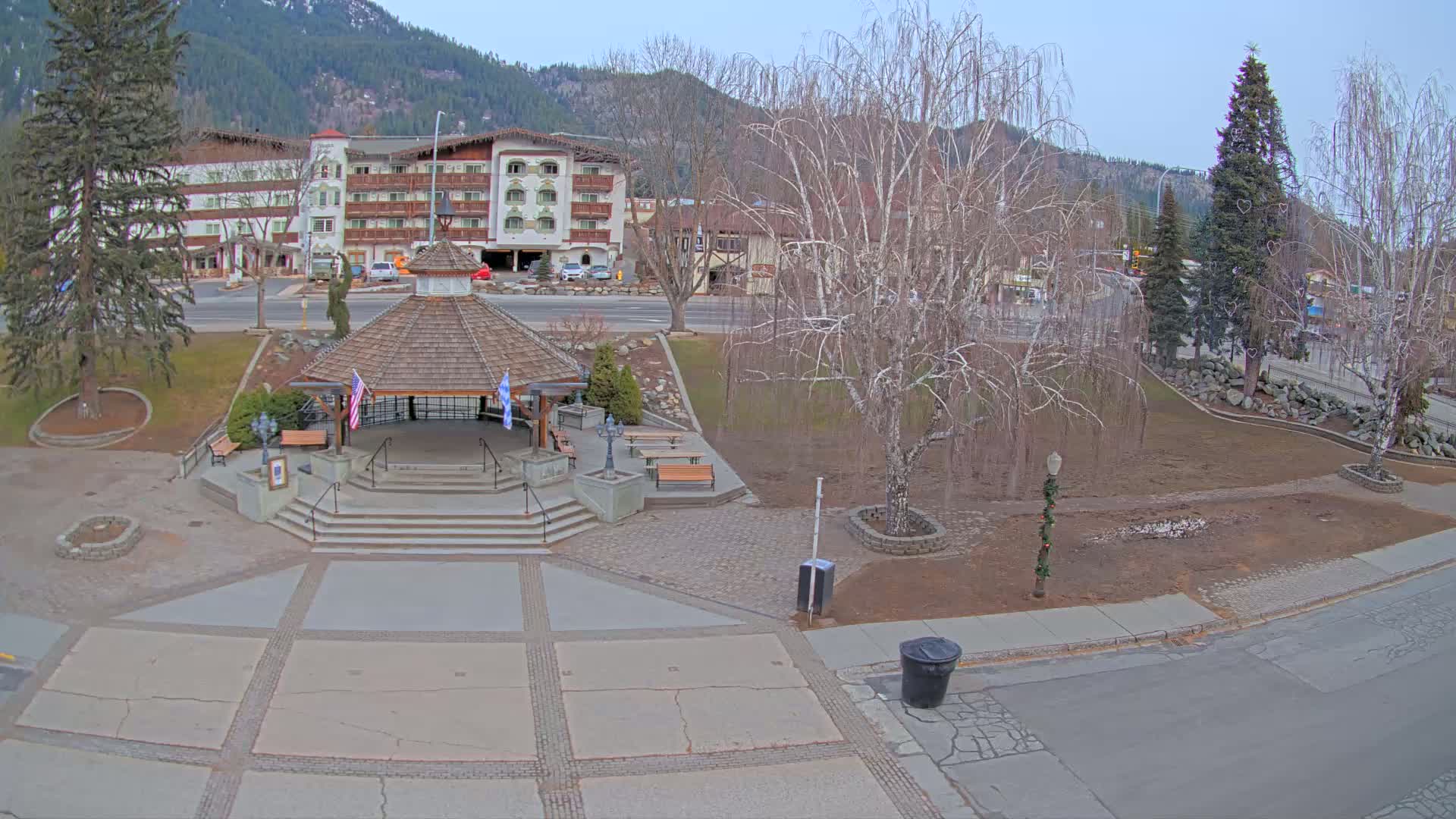 A paved town square with a gazebo, benches, and a bare birch tree sits in front of a multi-story building against a backdrop of mountains under a clear, twilight sky.