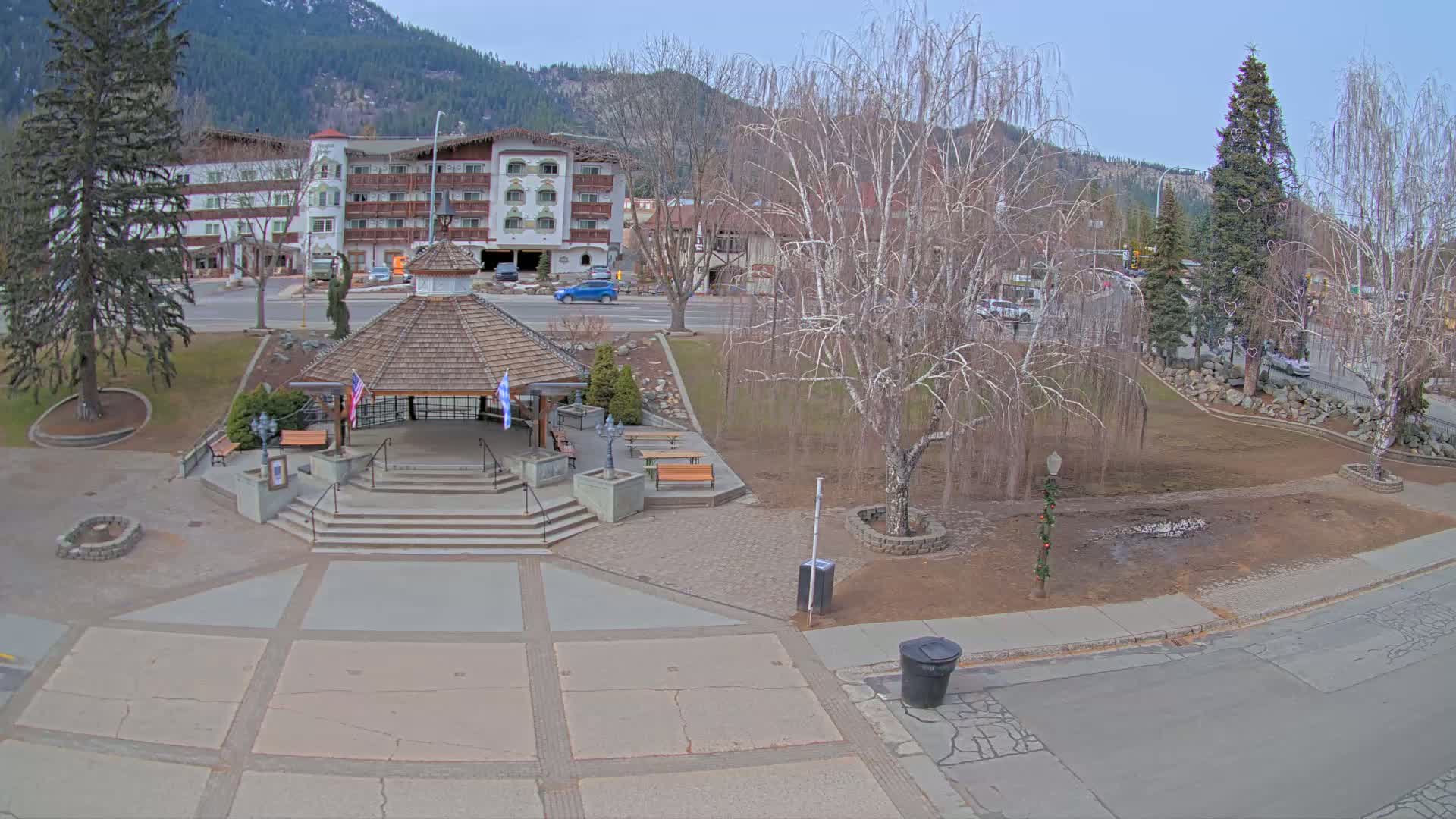 A gazebo with flags sits in a town square next to a multi-story building, under a mostly clear sky.