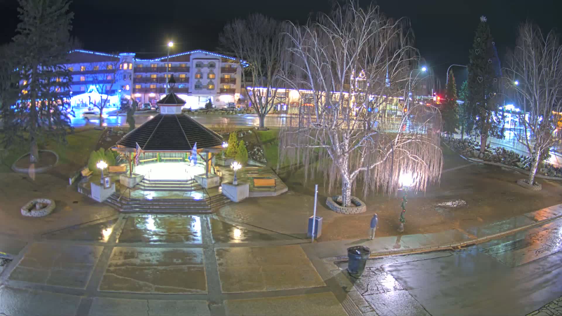 A nighttime view of a town square with a gazebo, wet pavement, bare trees, and a multi-story building in the background under a rainy sky.