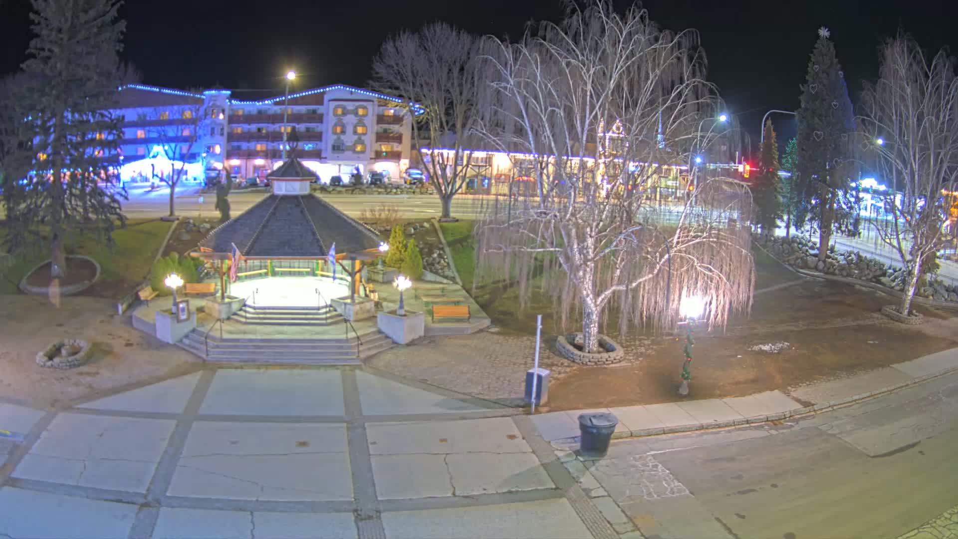 A nighttime view of a town square with a gazebo, illuminated trees, and a multi-story building in the background.
