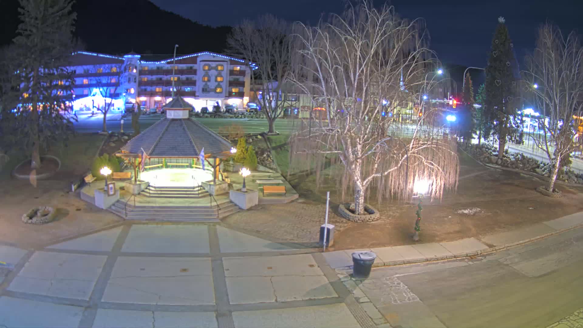 A nighttime view of a town square with a gazebo, a multi-story building, bare trees, and a decorated Christmas tree.