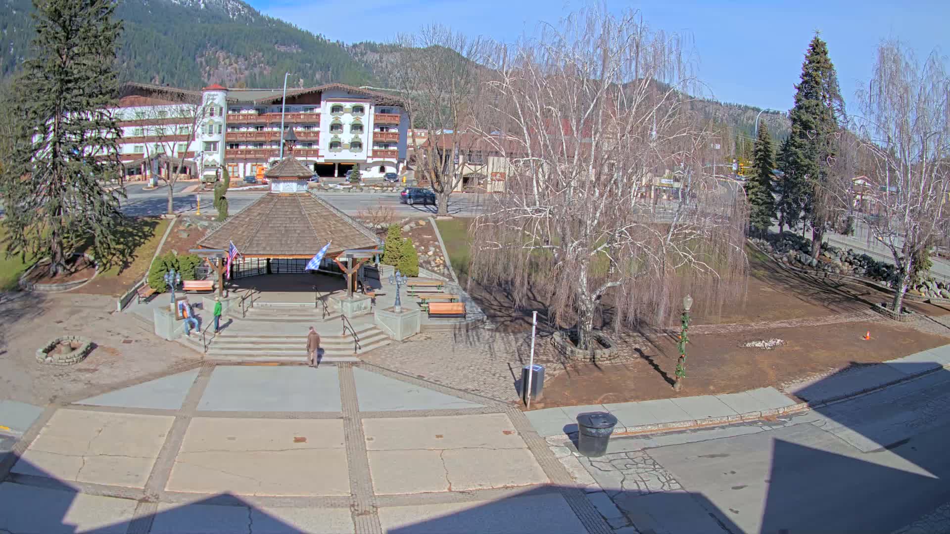 A sunny day reveals a town square with a gazebo, several people, bare trees, and a multi-story building in the background, all set against a backdrop of mountains.