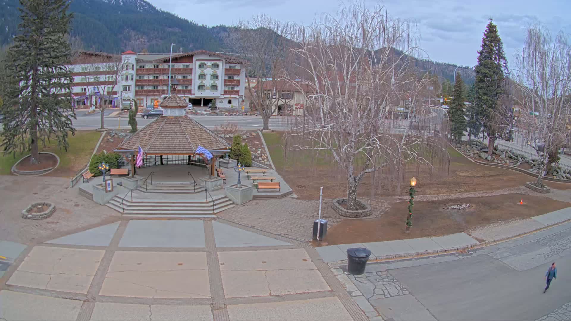 A gazebo sits in a town square with a paved area, a few benches, trees, and a multi-story building in the background under a cloudy sky.