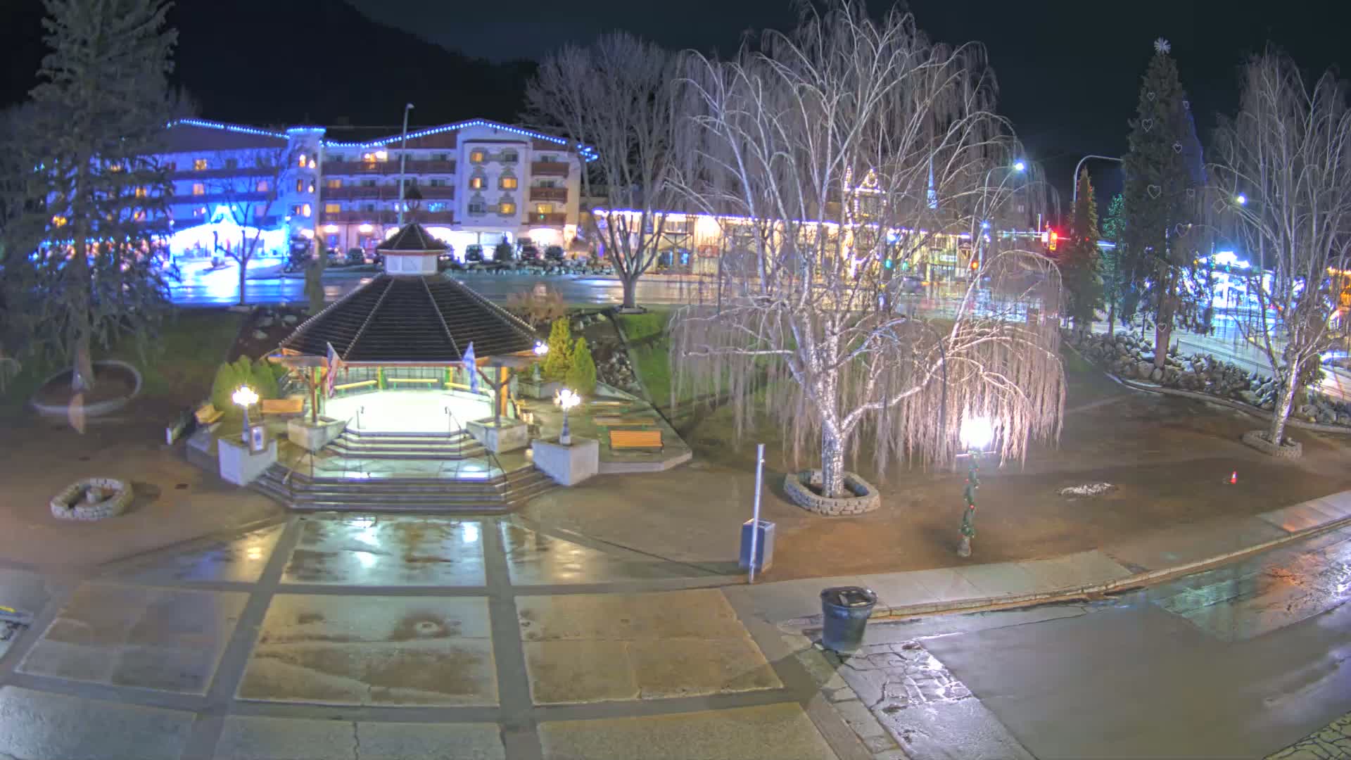 A nighttime view of a town square with a gazebo, trees, and a multi-story building, all wet from recent rain or snow.