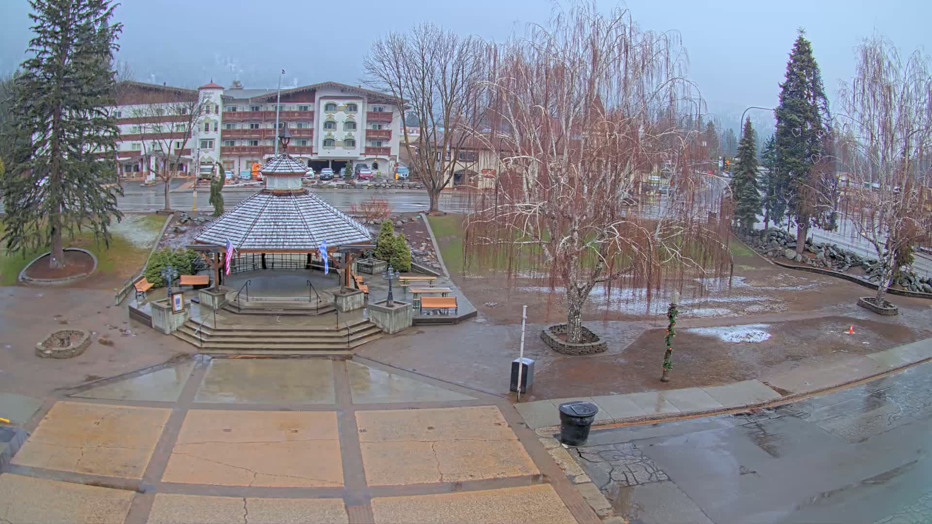 A snow-dusted gazebo sits in a town square, surrounded by bare trees and wet pavement under an overcast sky.