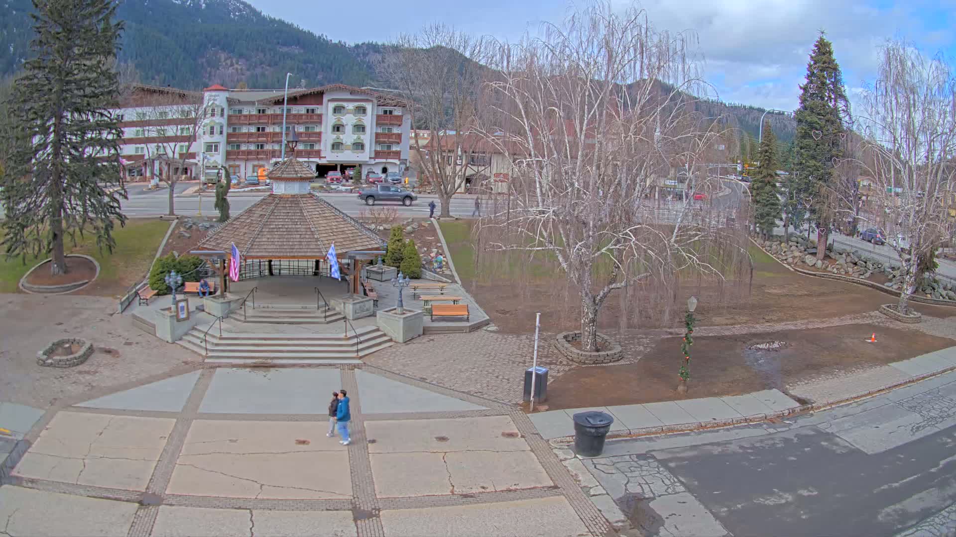 A town square with a gazebo, benches, and a couple walking, situated in front of a hotel and mountains under a partly cloudy sky.
