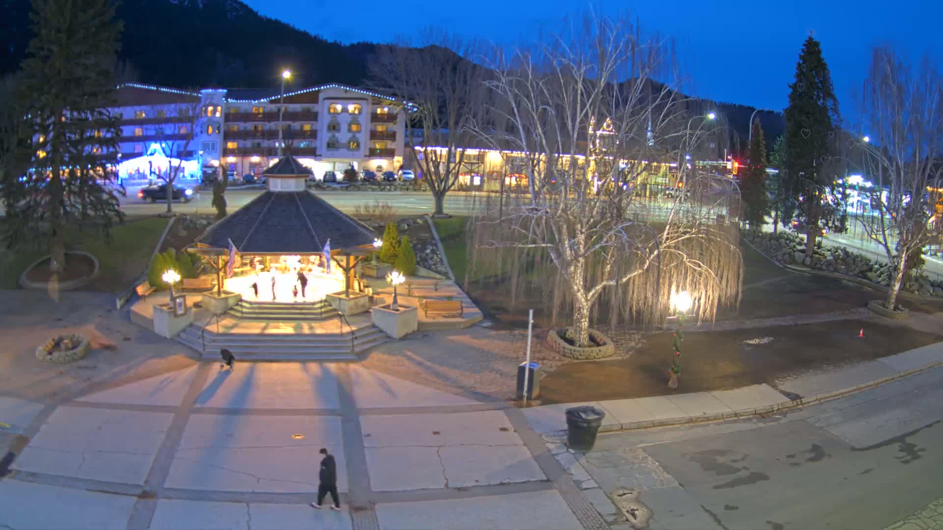A nighttime view of a town square with a gazebo, illuminated buildings, and bare trees under a twilight sky.