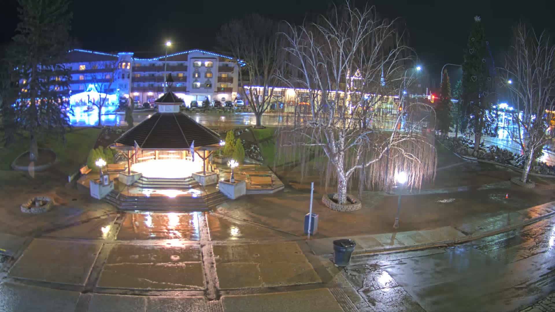 A nighttime view of a wet town square featuring a gazebo, benches, and bare trees under lights, with a building and a decorated Christmas tree in the background.
