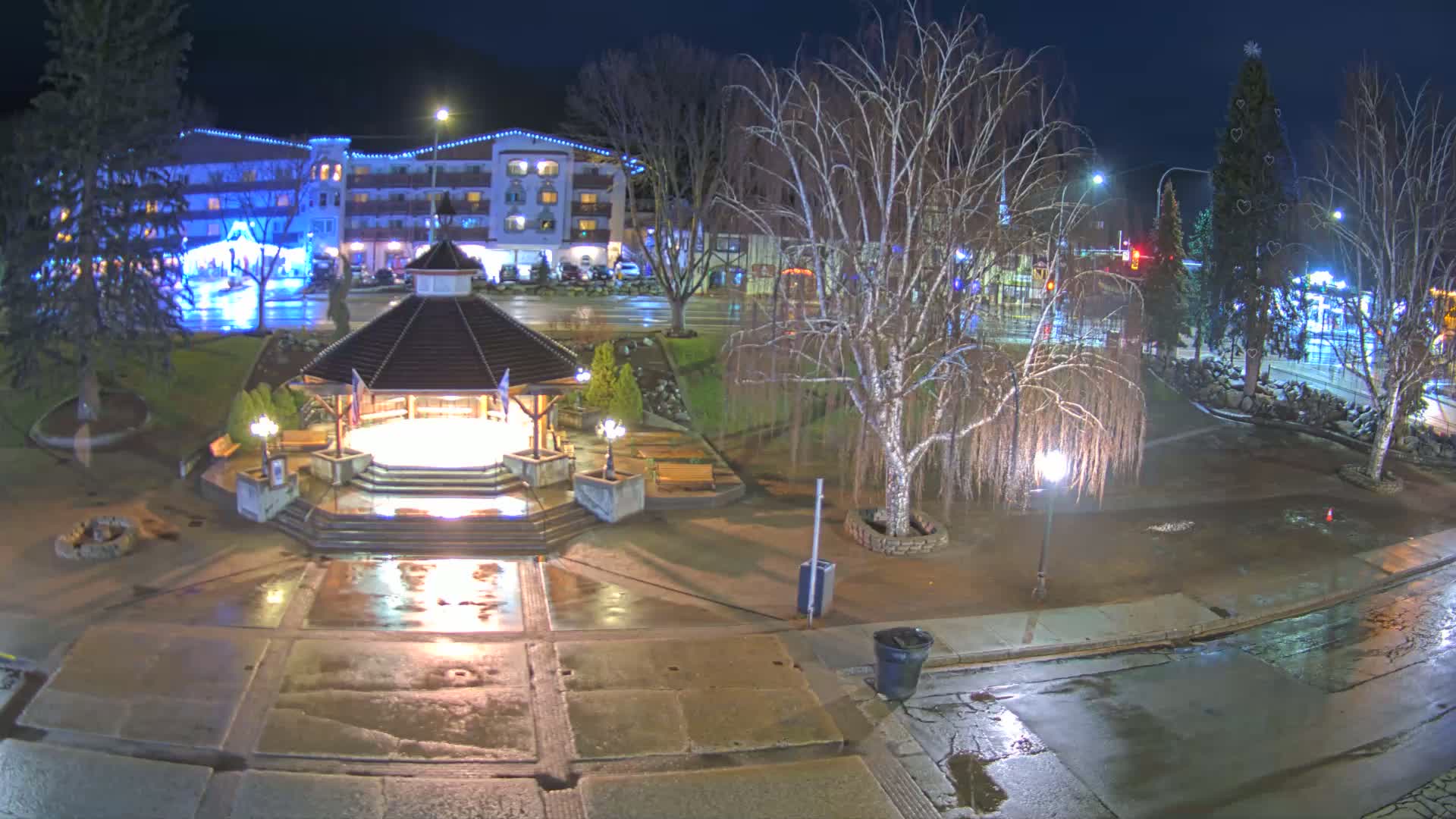 A nighttime view of a town square, featuring a gazebo, benches, and trees, with wet pavement indicating recent rain.
