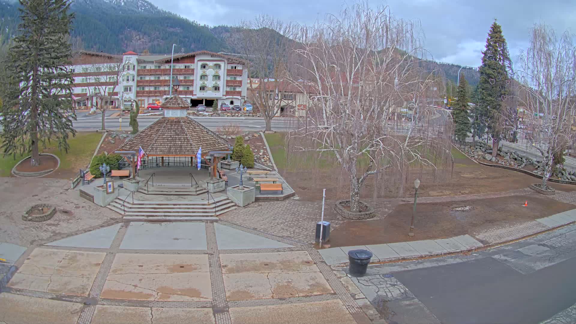 A paved town square with a gazebo, benches, and bare trees sits in front of a multi-story building against a backdrop of mountains under an overcast sky.