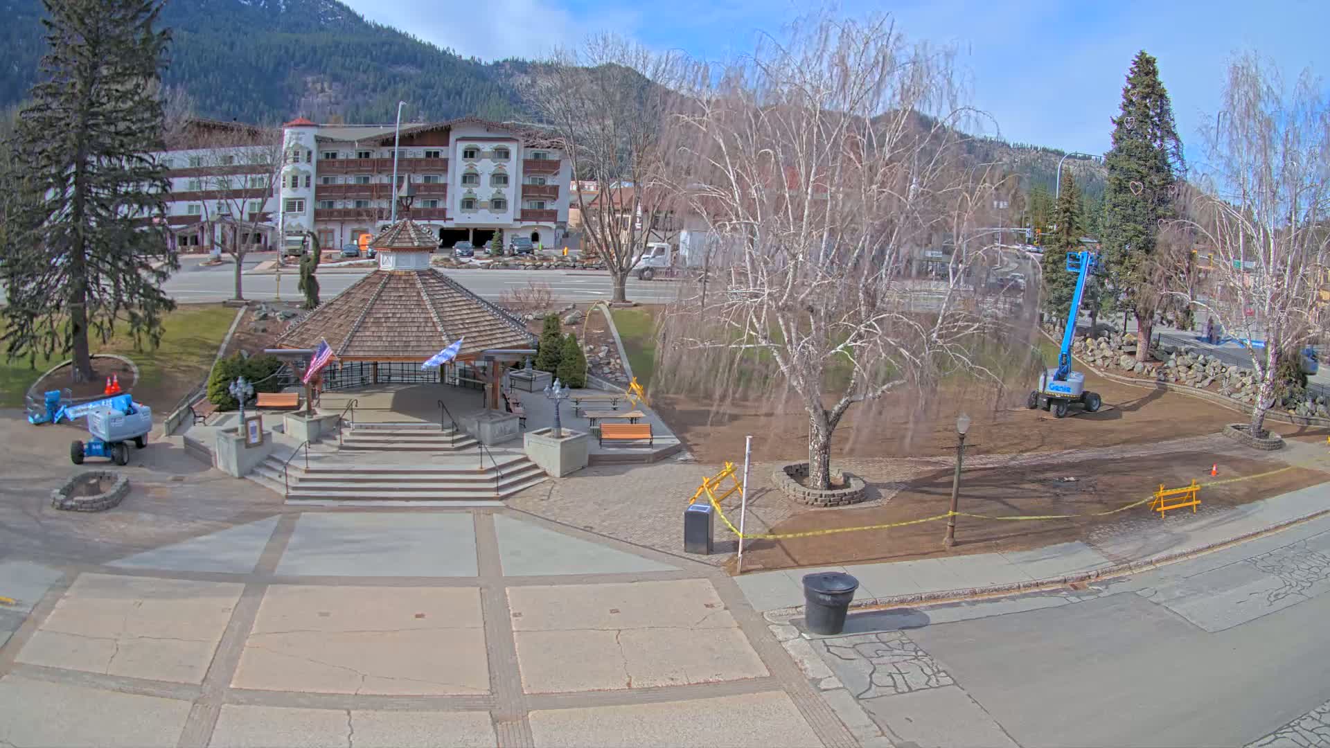 A town square with a gazebo, benches, and landscaping under a mostly sunny sky, with a mountain range in the background.