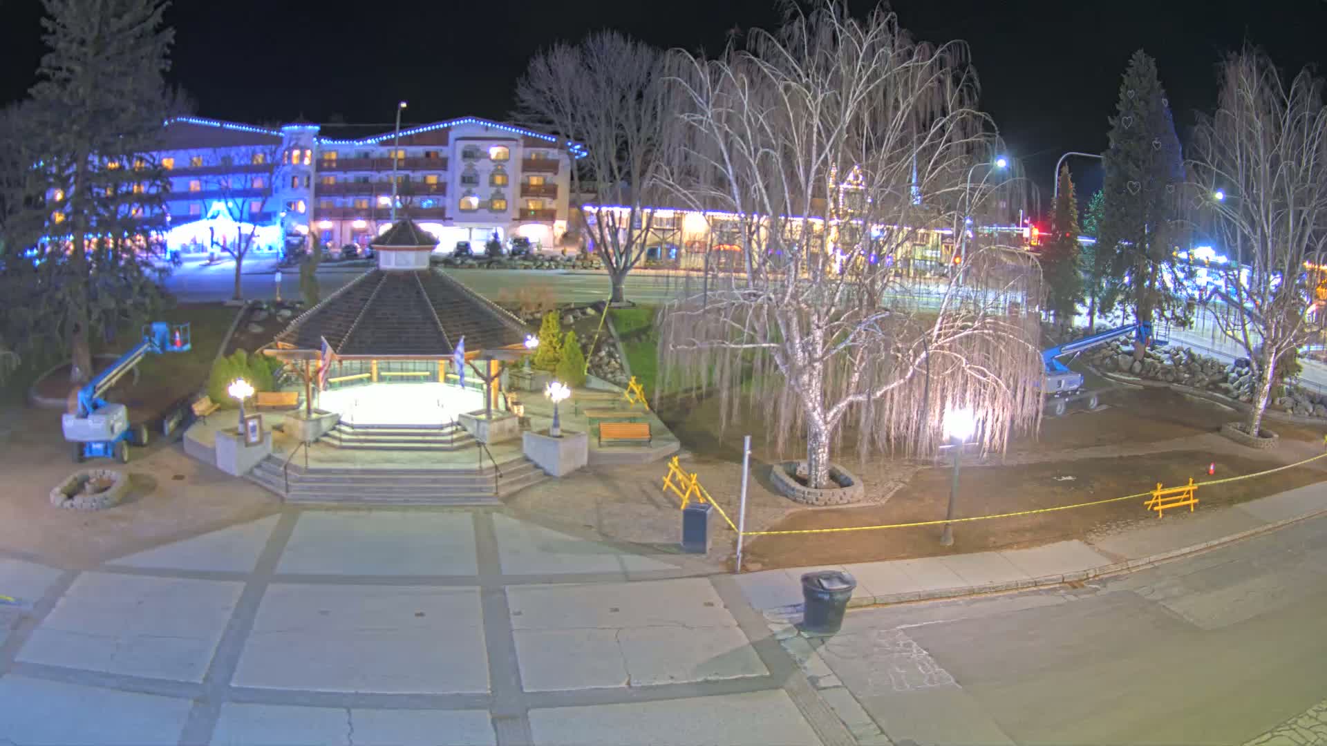 A nighttime view of a town square with a gazebo, bare trees, and a multi-story building illuminated with lights.