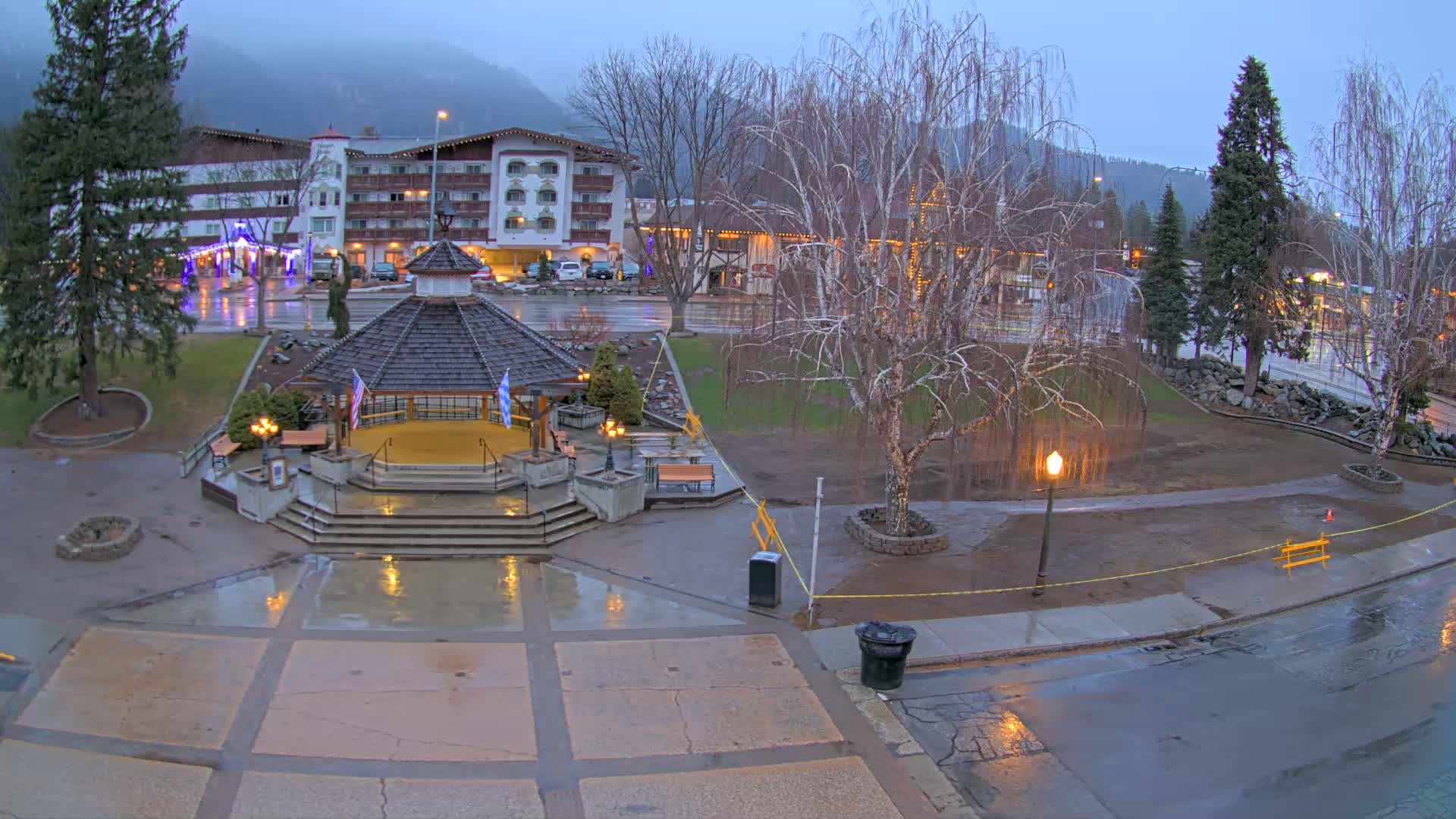 A wet town square with a gazebo in the center, situated near a multi-story hotel under a gloomy sky.