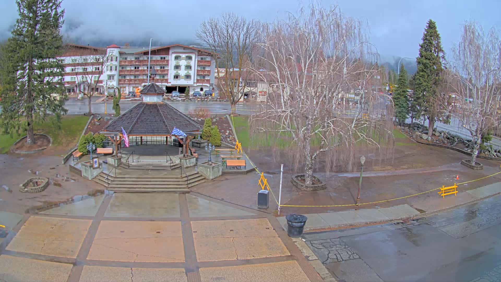 A wet town square with a gazebo in the center, surrounded by bare trees and a multi-story building, under an overcast sky.