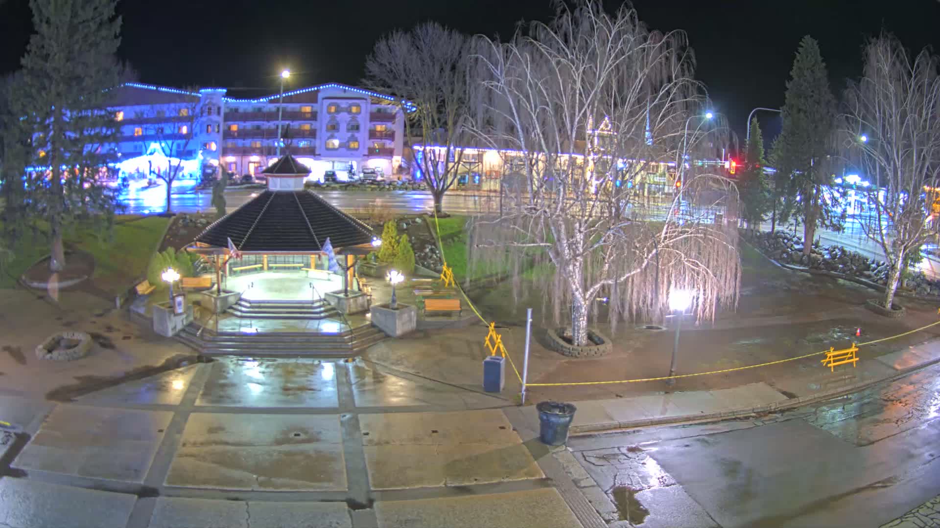A nighttime view of a wet town square with a gazebo, trees, and a hotel in the background under partly cloudy skies.