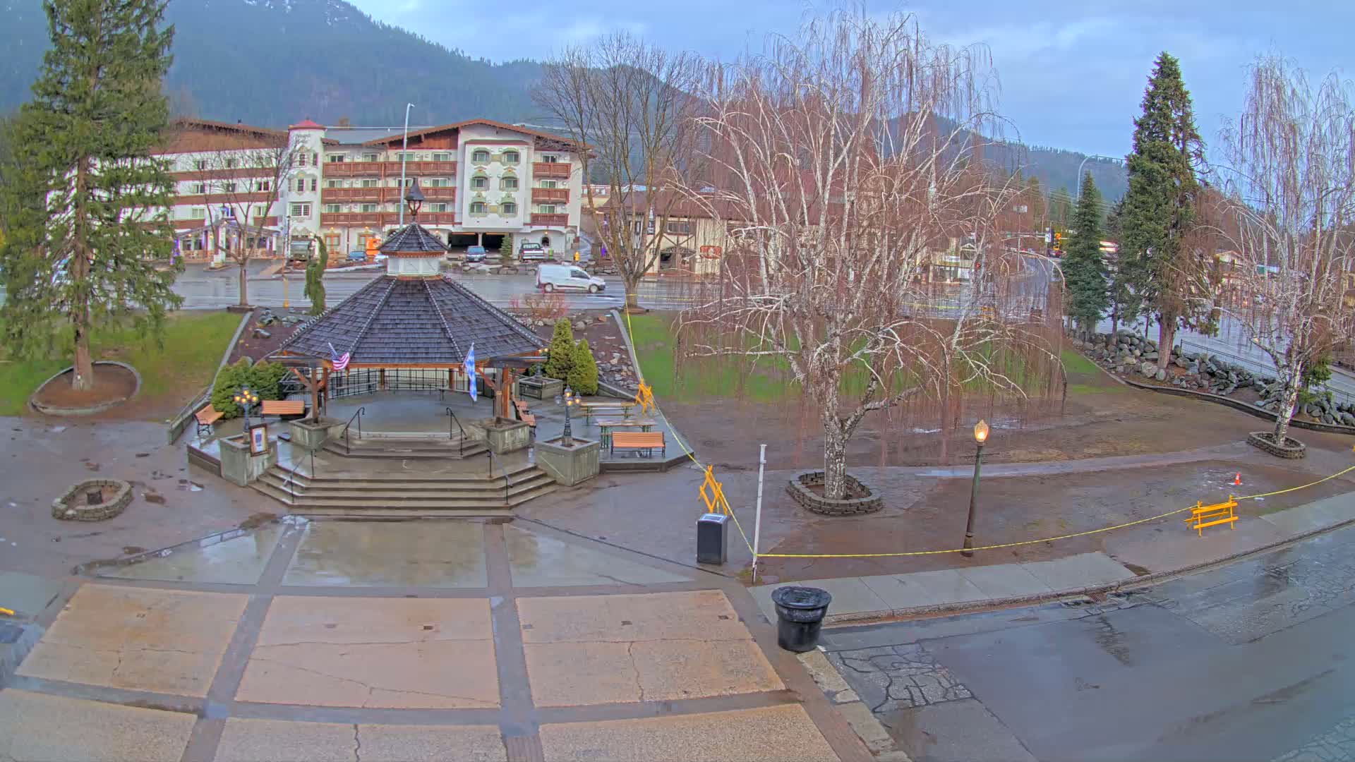 A wet town square with a gazebo, benches, and a multi-story building in the background under an overcast sky.