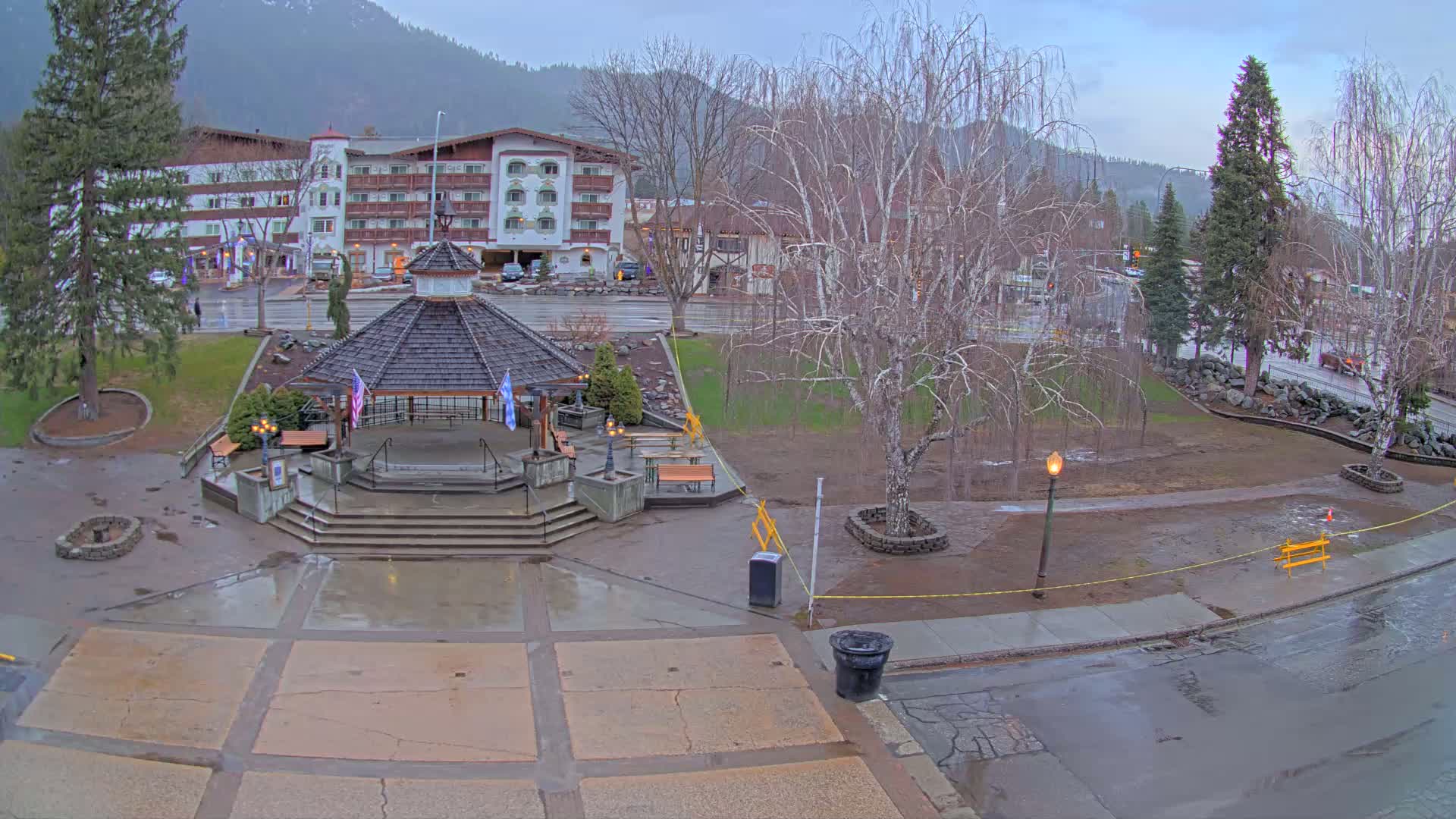 A wet town square with a gazebo in the center, surrounded by trees, a hotel, and a street under an overcast sky.