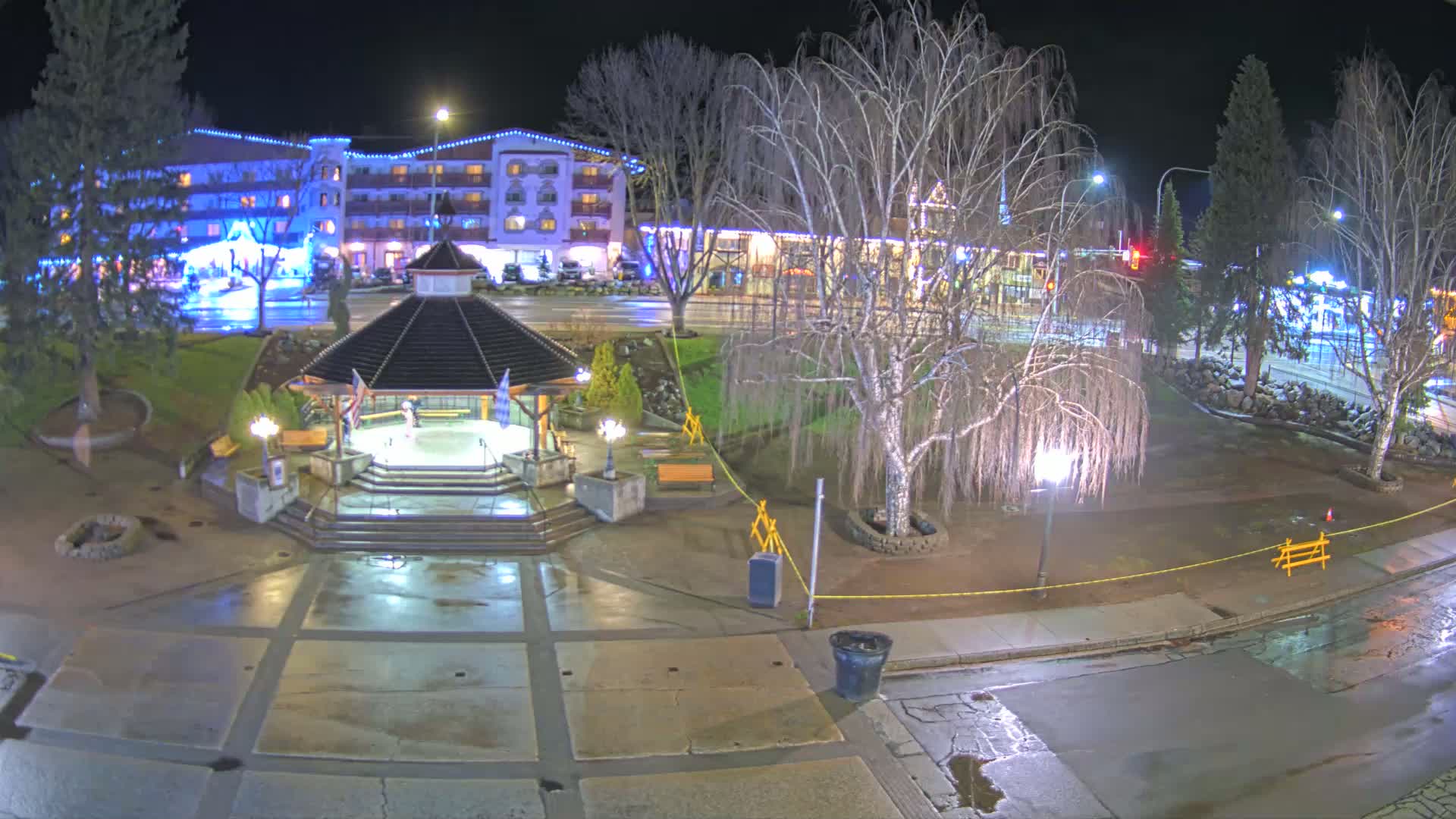 A nighttime view of a town square with a gazebo, trees, a multi-story building, and wet pavement.