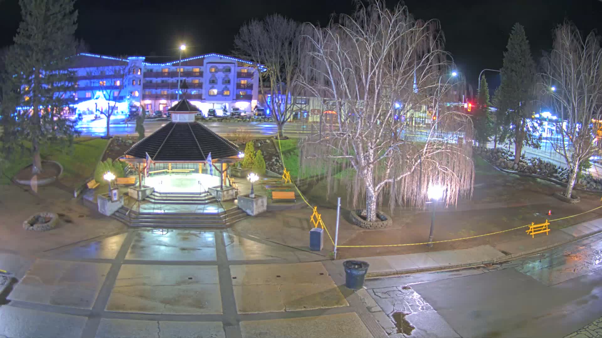 A nighttime view of a town square with a gazebo, a multi-story hotel in the background, and wet pavement under partly cloudy skies.