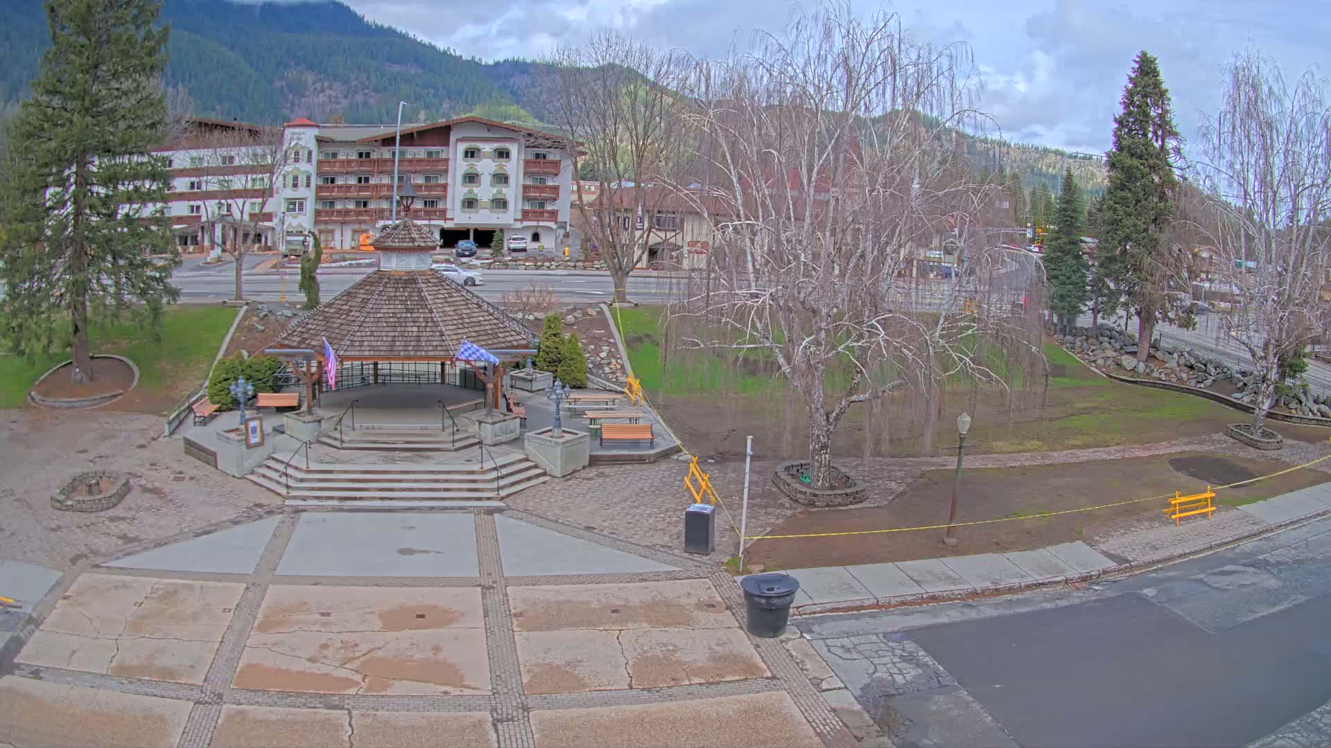 A park with a gazebo and benches sits in front of a hotel and mountain range under a cloudy sky.