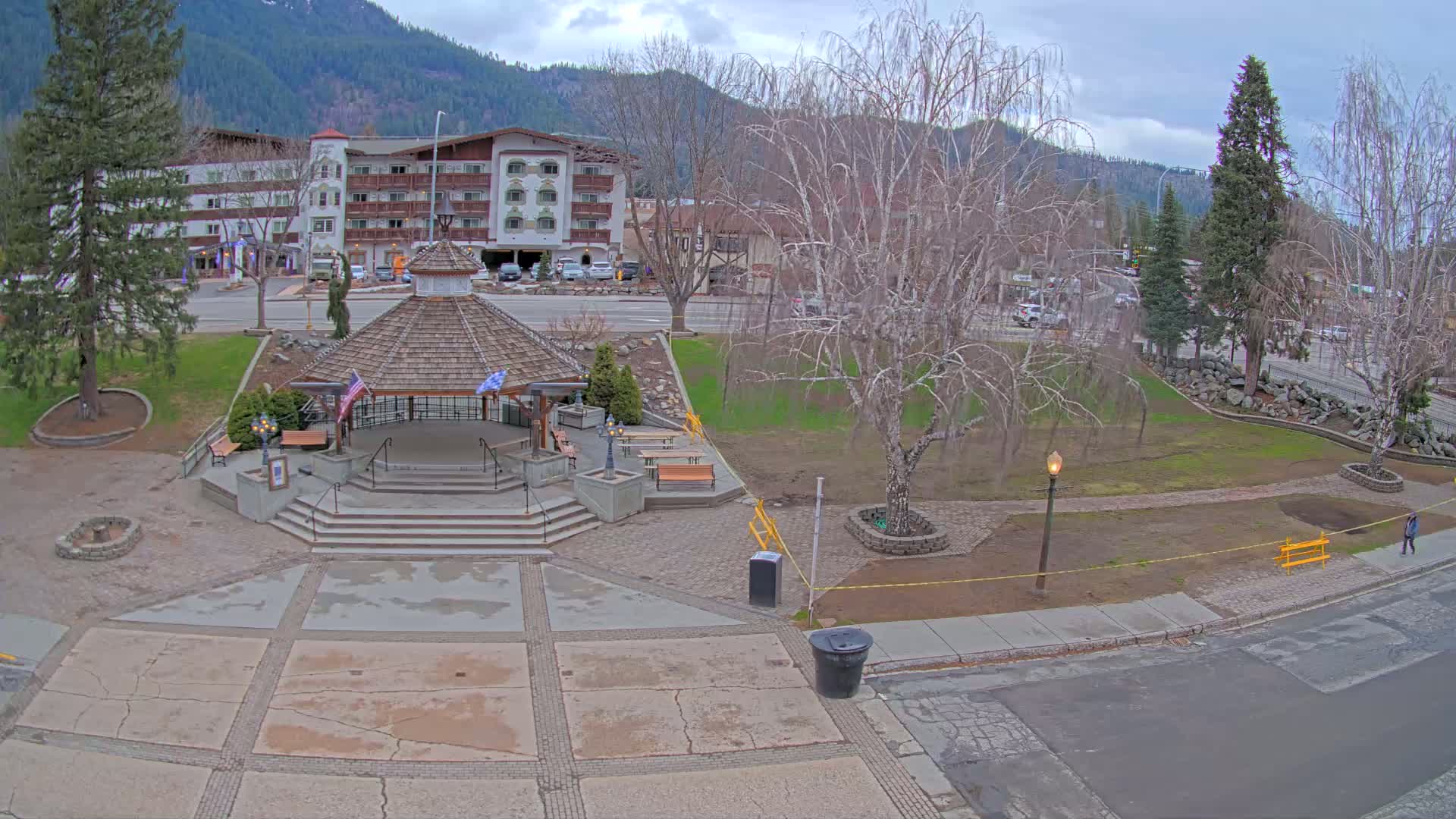 A park with a gazebo and several benches sits in front of a multi-story building, all under a cloudy sky in a mountainous region.
