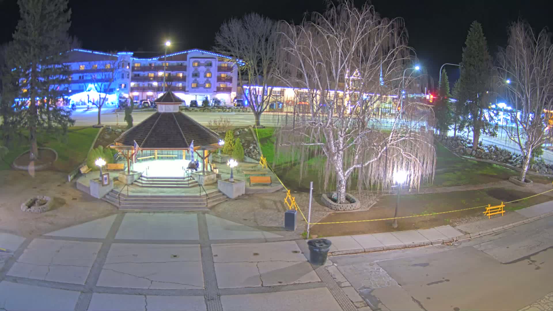 A nighttime view of a town square featuring a gazebo, bare trees, and a multi-story building illuminated with string lights.