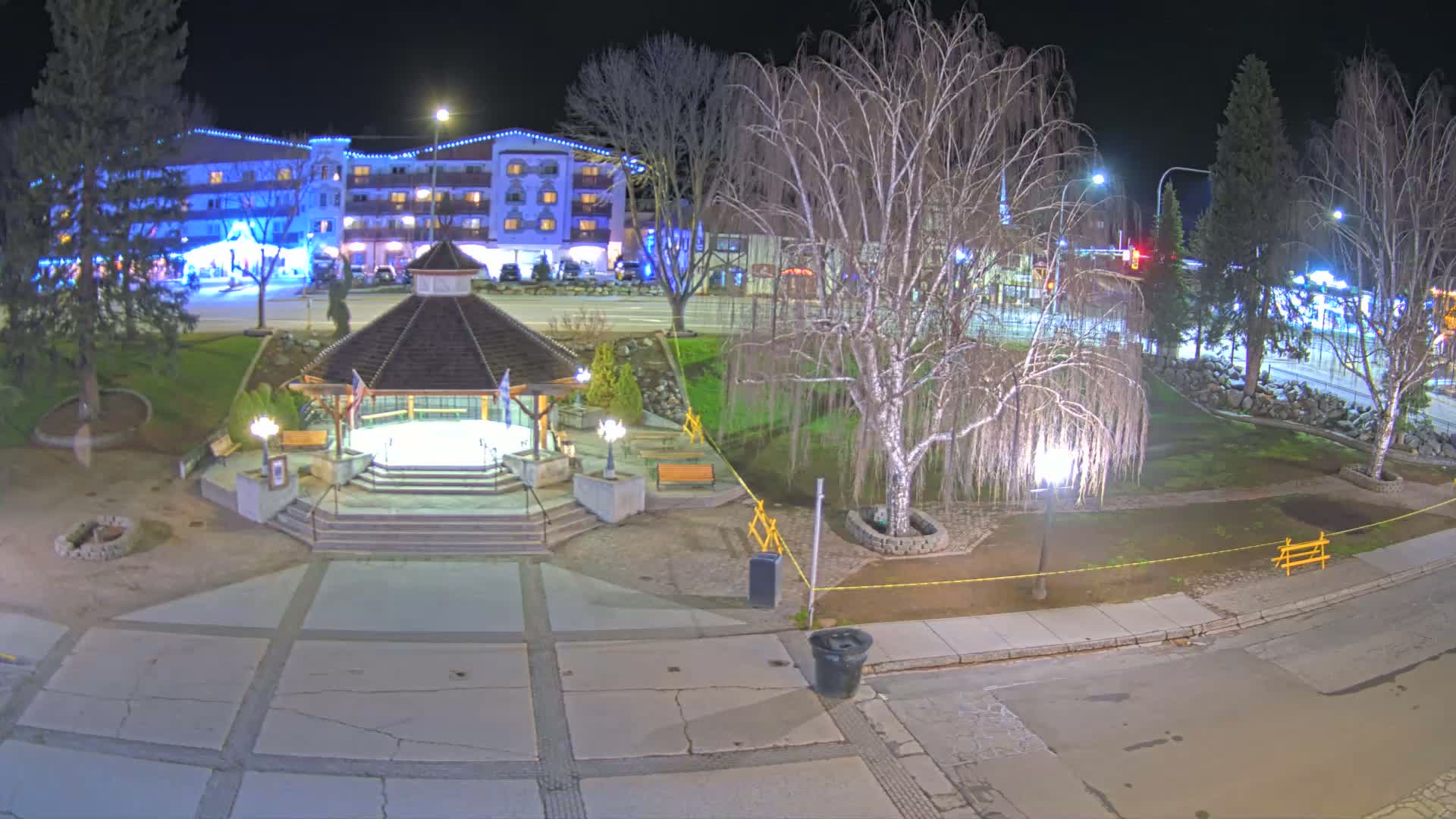A nighttime view of a town square with a gazebo, trees, and a multi-story building in the background.