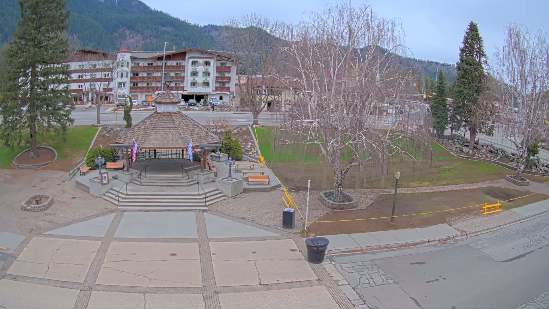 A park with a gazebo, benches, and a bare tree sits in front of a multi-story building, all under a cloudy sky.
