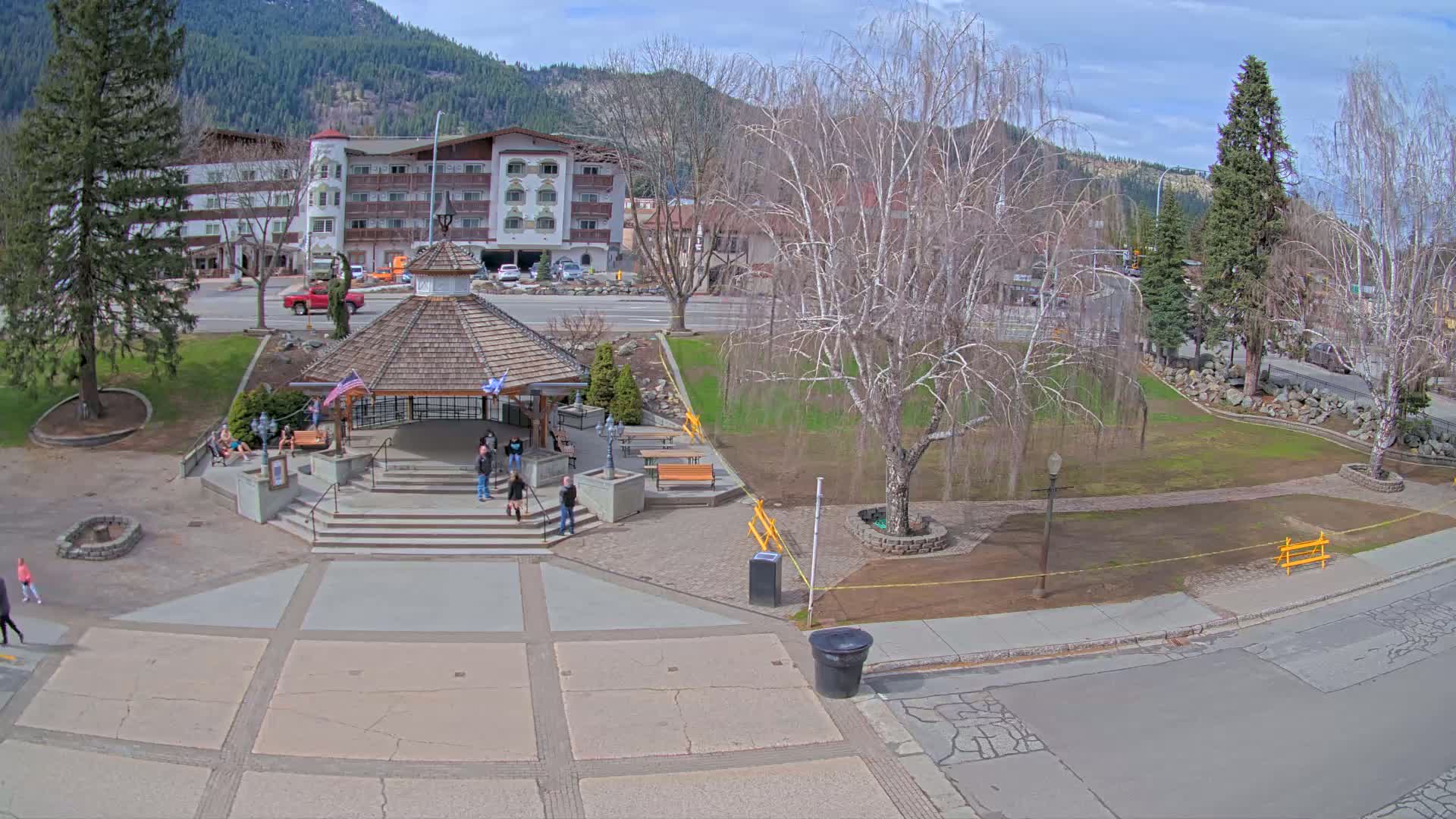 A town square with a gazebo and several people, situated in front of a multi-story building, under a partly sunny sky with mountains in the background.