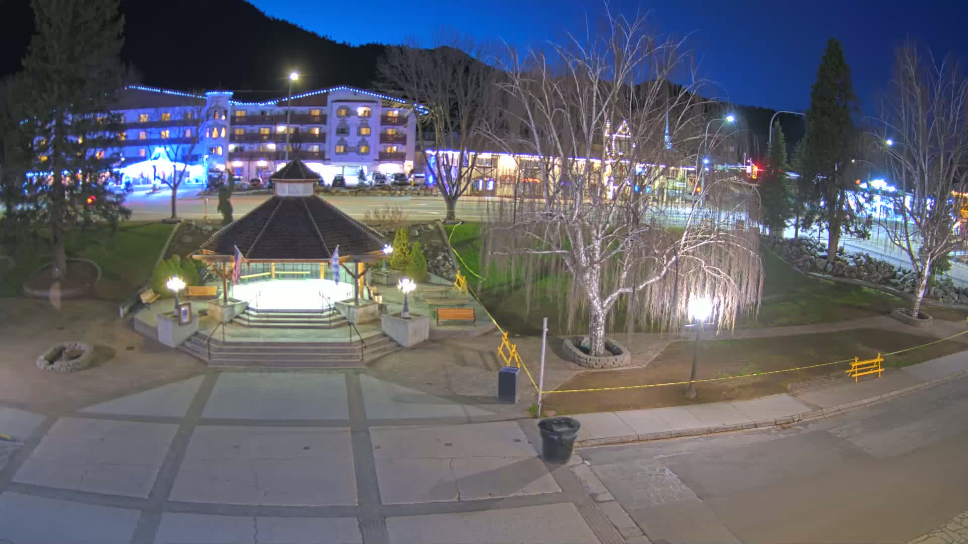 A nighttime view of a town square with a gazebo, illuminated buildings, and bare trees under a dark blue sky.