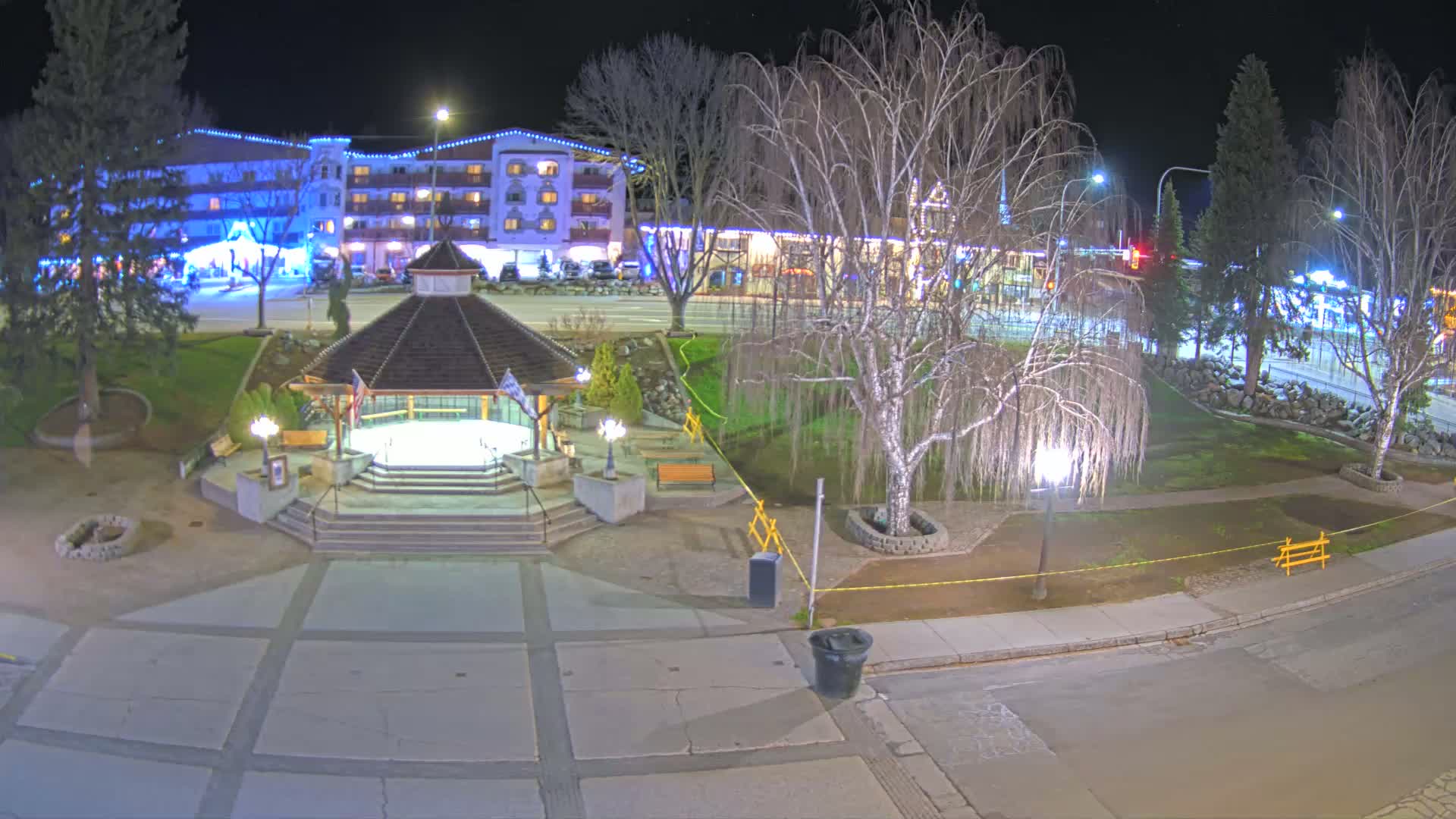 A nighttime view of a town square with a gazebo, trees, and a multi-story building in the background, under clear skies.