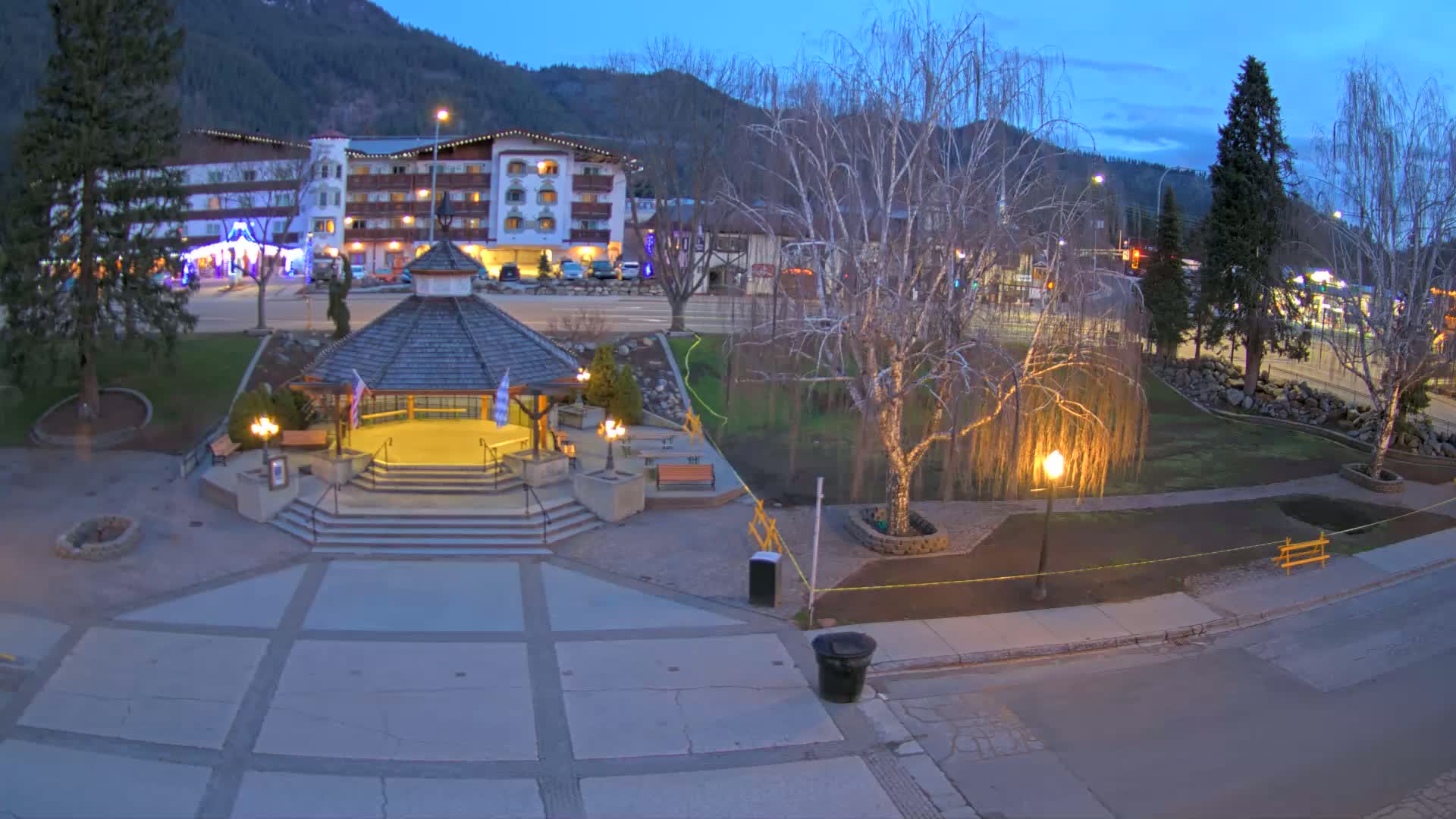 A gazebo sits in a town square at dusk, with a hotel and mountains in the background under a twilight sky.