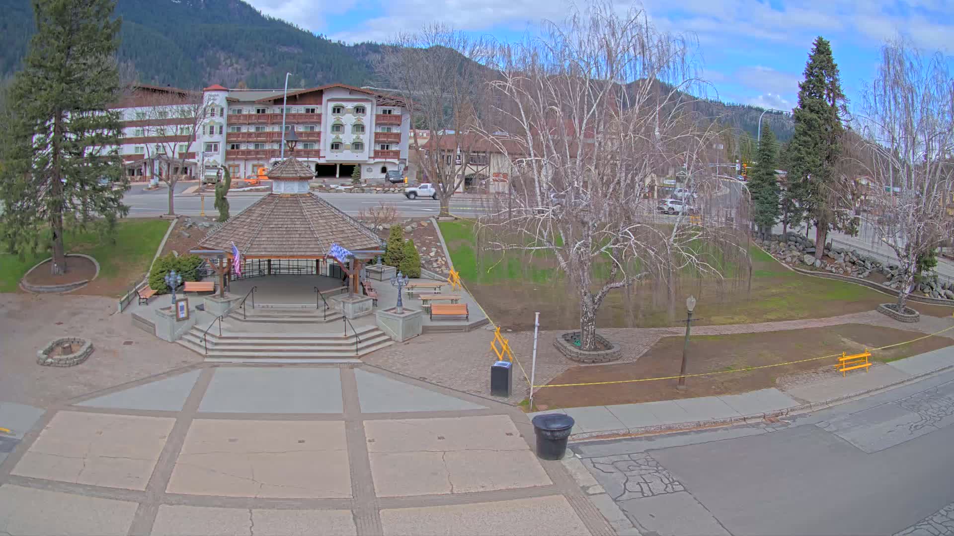 A gazebo sits in a town square, under a partly cloudy sky, with a hotel and mountains in the background.