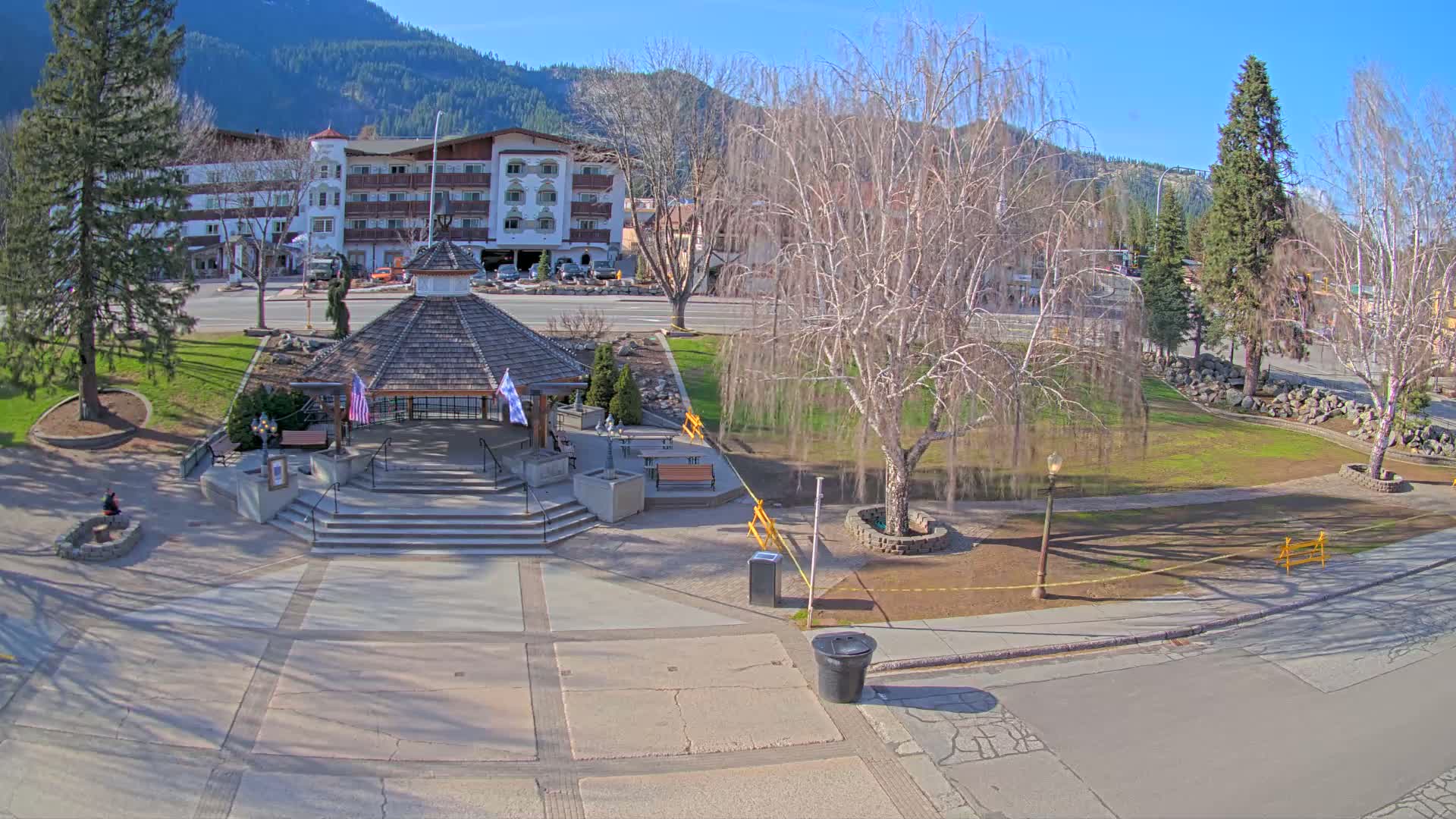 A gazebo sits in a paved plaza next to a grassy park, with a multi-story building and mountains in the background on a sunny day.