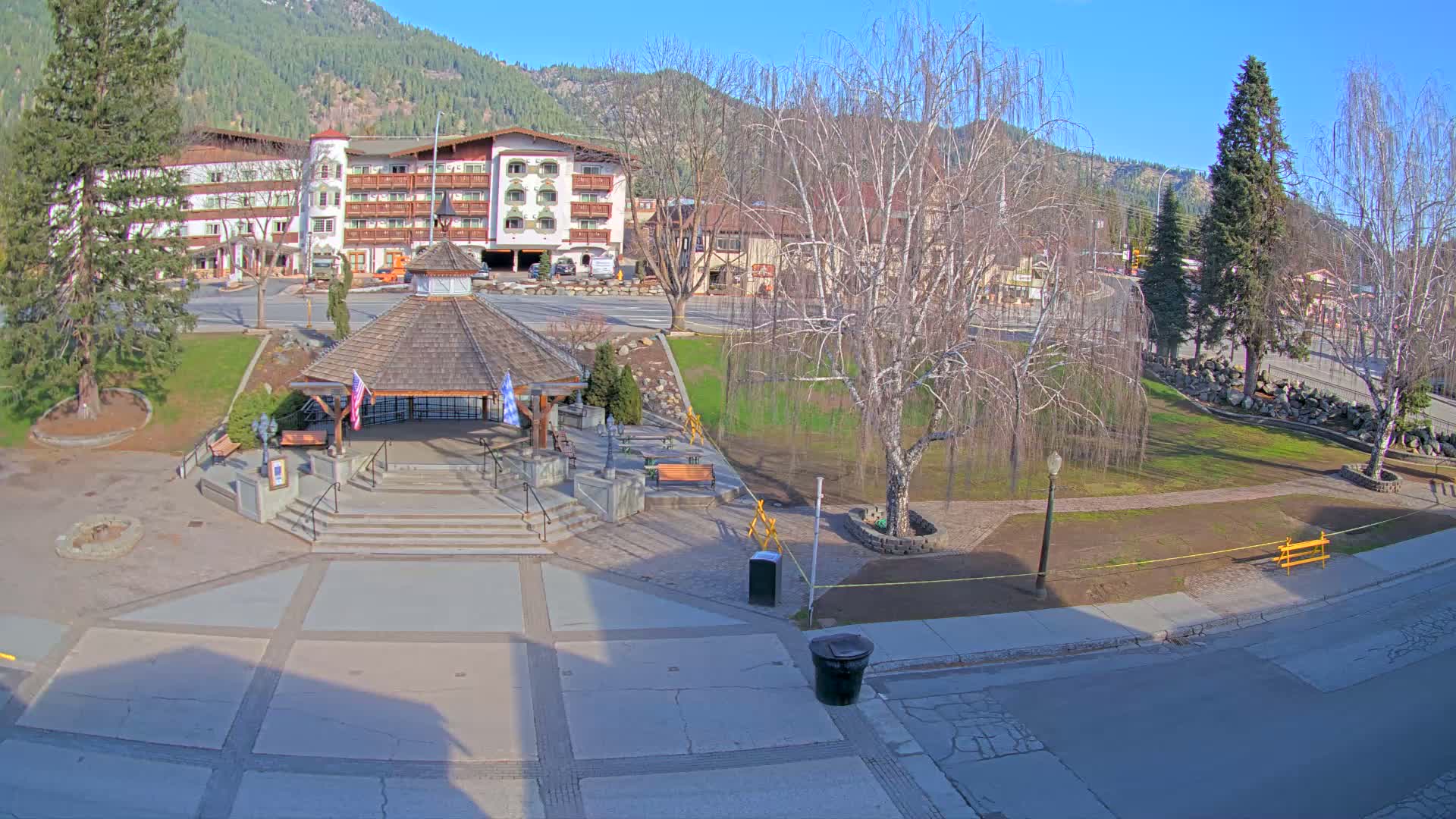 A town square with a gazebo, benches, and a multi-story building in the background, under a sunny sky.