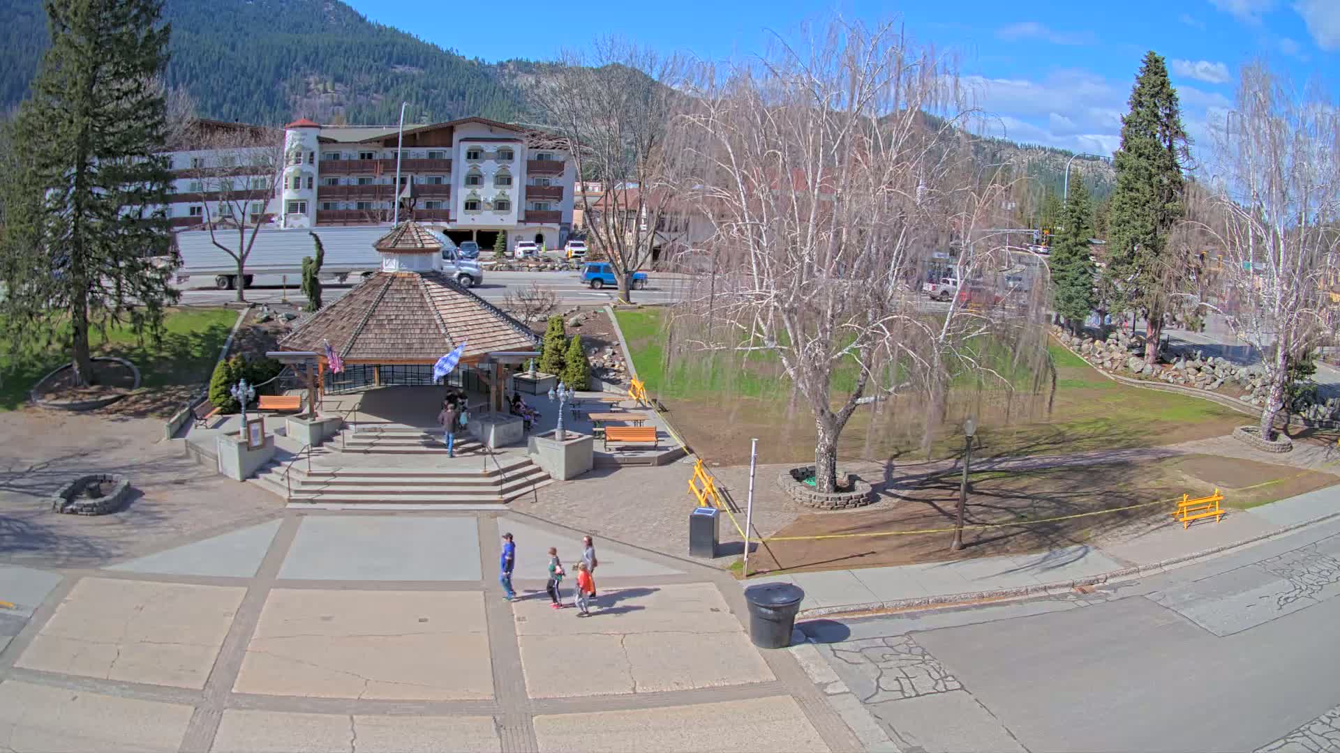 A paved plaza with a gazebo and a few people, situated in front of a multi-story building, under a sunny sky.