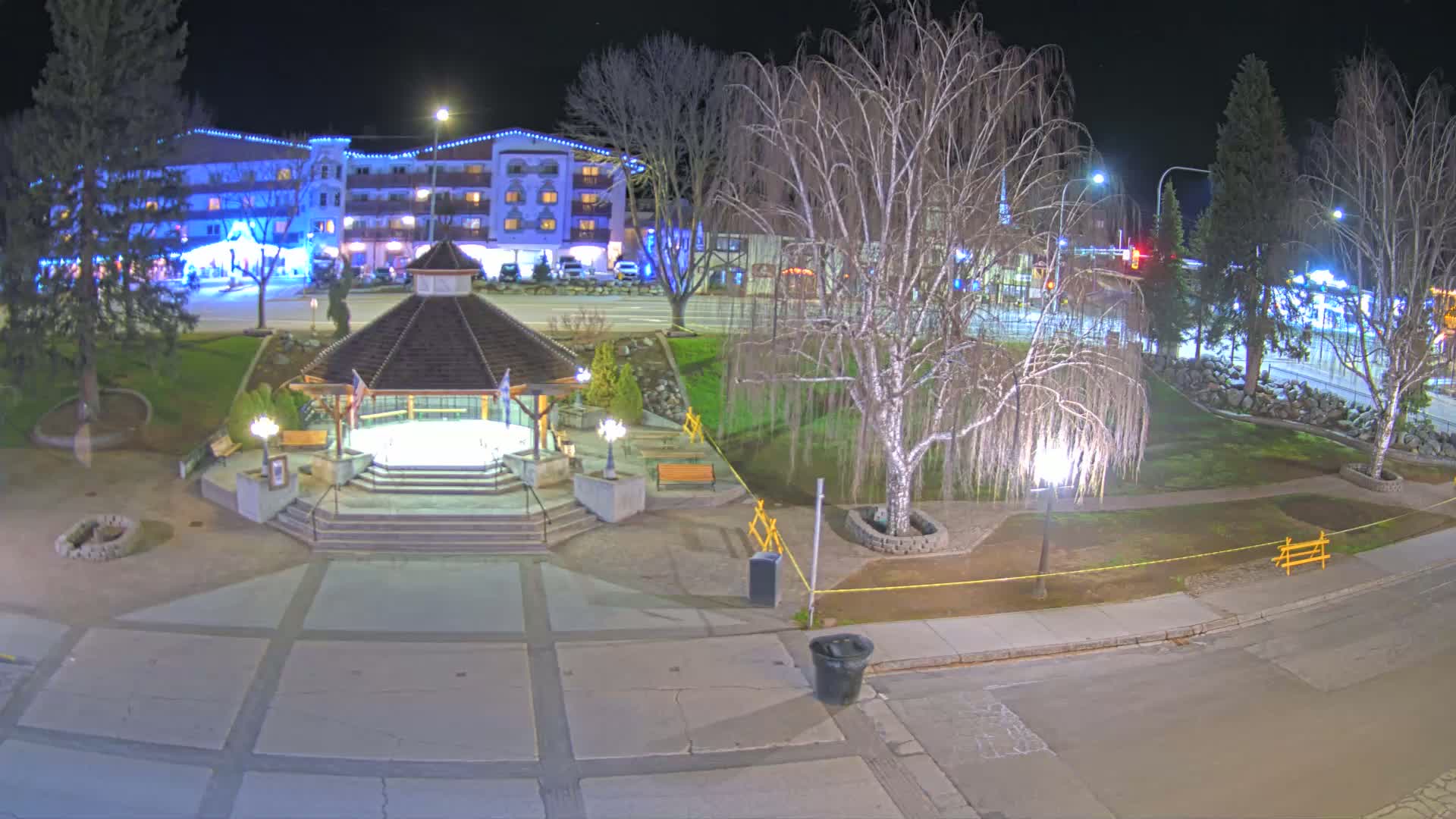 A nighttime view of a town square with a gazebo,  trees, and a multi-story building in the background under clear skies.