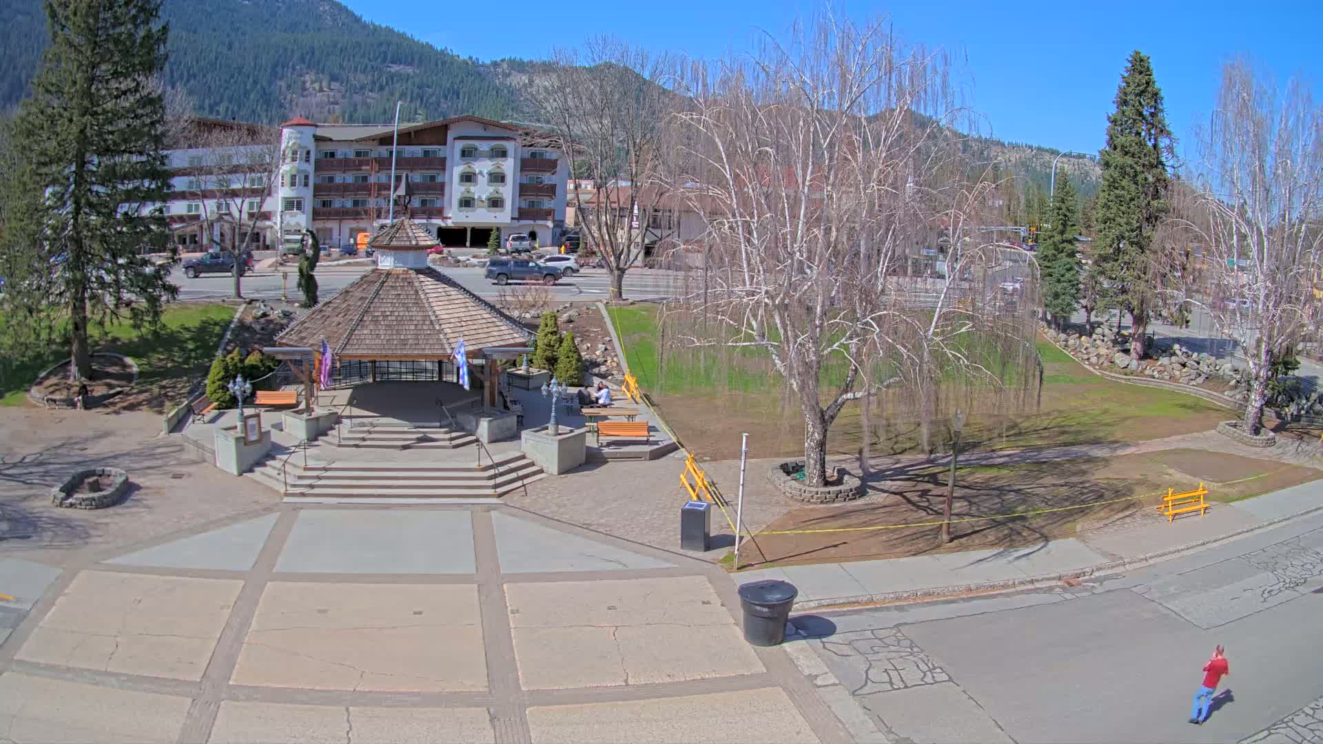 A paved plaza with a gazebo, benches, and trees, situated in front of a multi-story building against a backdrop of mountains under a sunny sky.