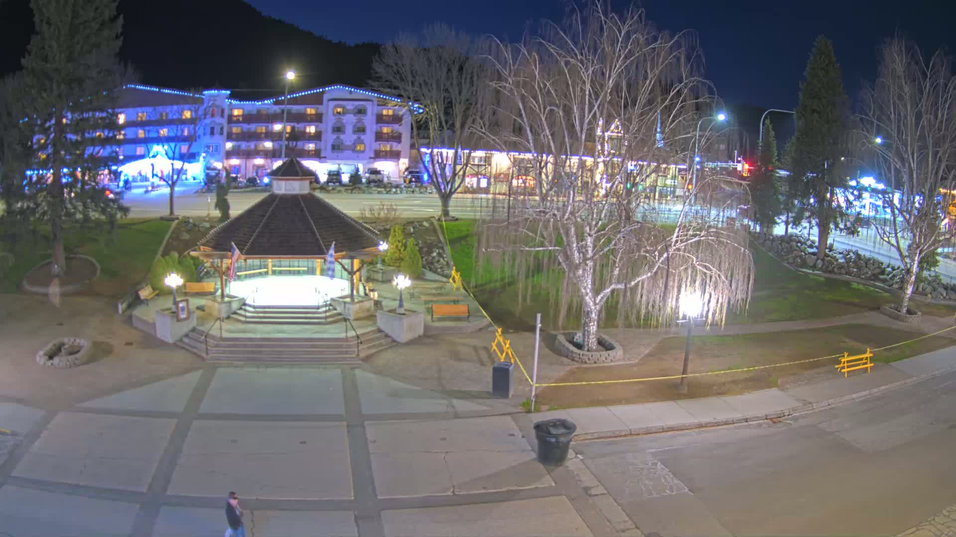 A nighttime view of a town square with a gazebo, illuminated buildings, and bare trees under a clear, dark sky.