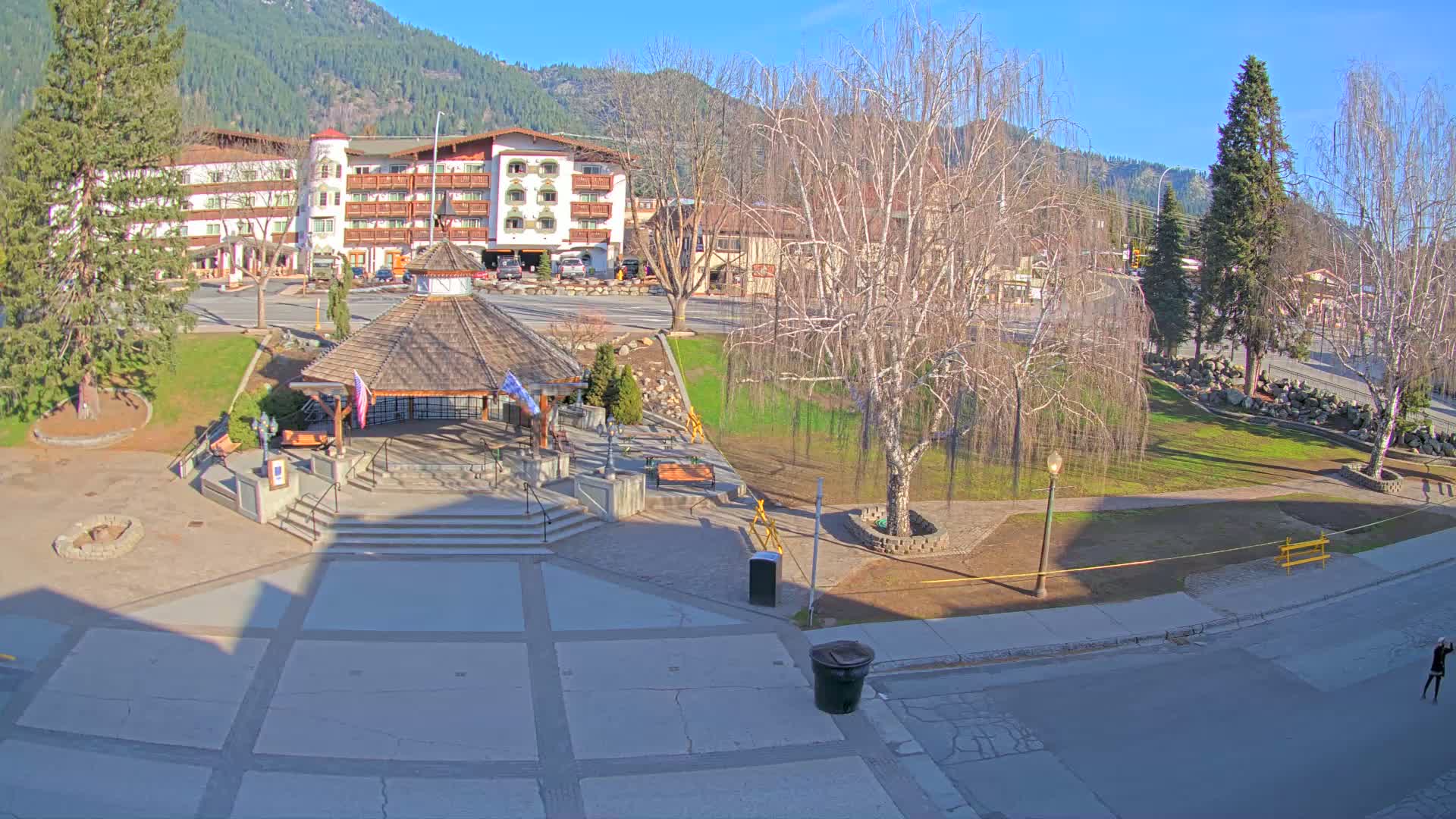 A town square with a gazebo, surrounded by trees, a building, and a paved area, on a sunny day with mountains in the background.