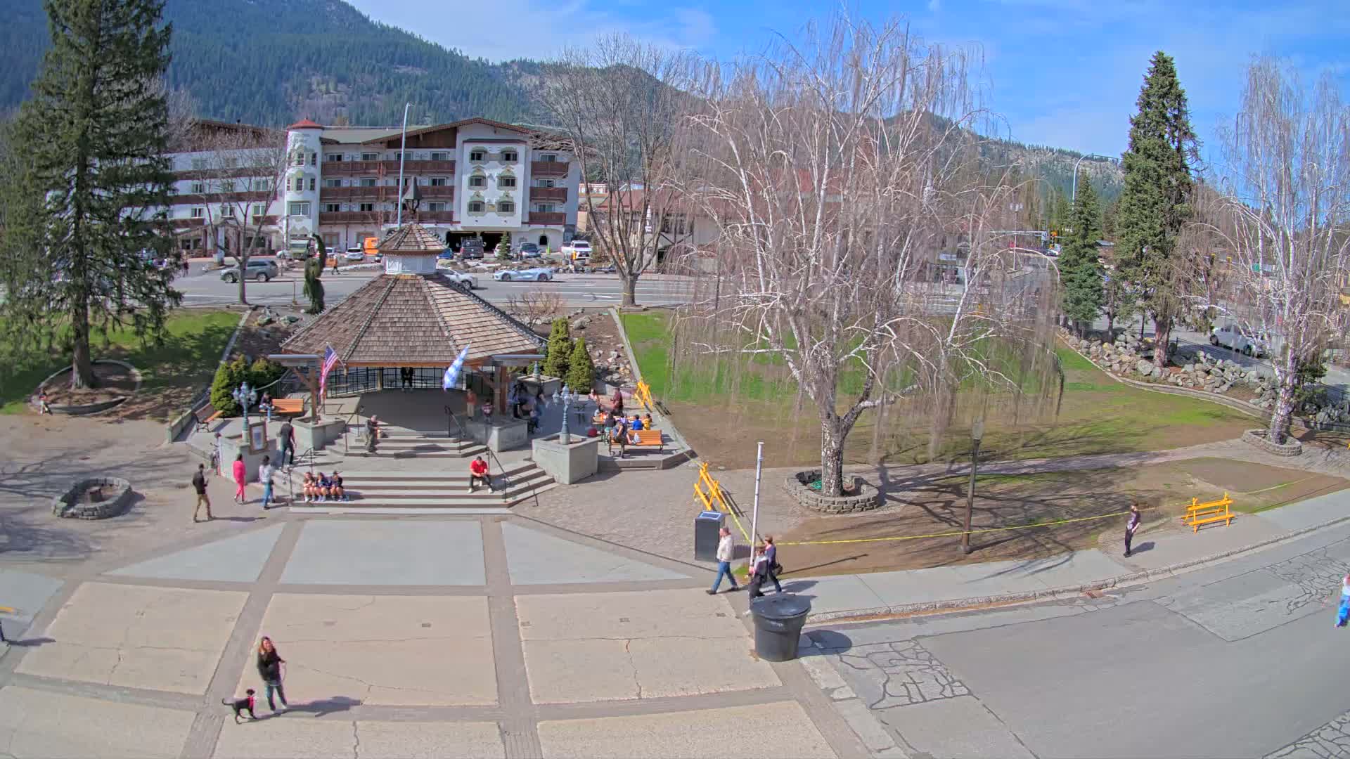 A sunny day in a town square with a gazebo, people walking around, and a multi-story building in the background.