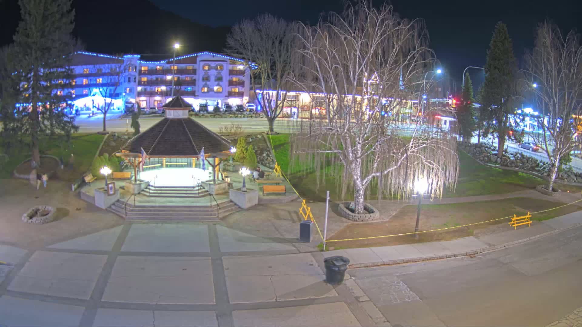 A nighttime view of a town square with a gazebo, trees, a hotel, and a street, illuminated by streetlights.